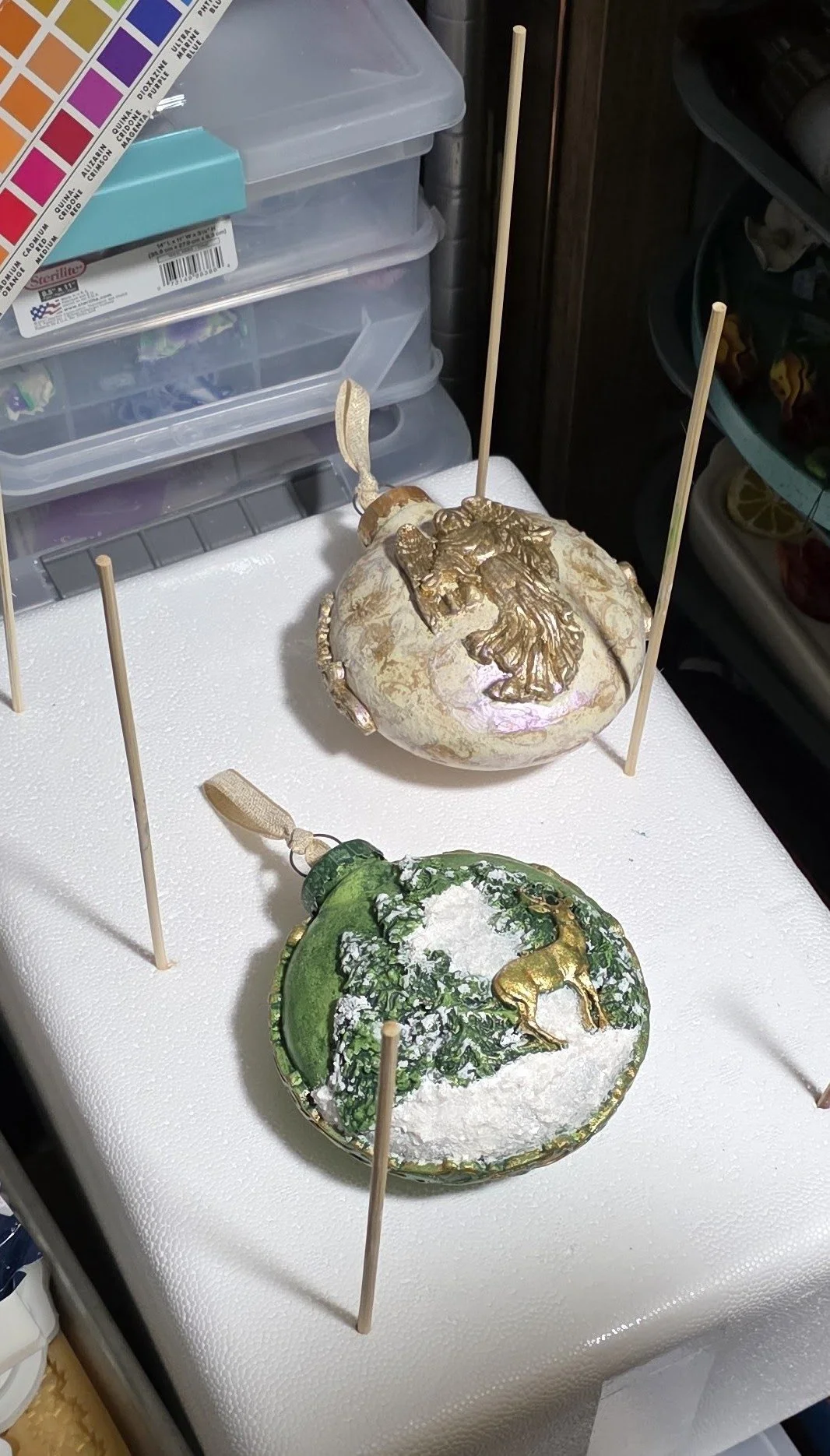 In our studio, a finished hand-sculpted and molded Guardian Angel Ornament by Art Illusions by Young sits beside a green Christmas tree ornament still in production. The Guardian Angel features soft iridescent pearl, blush, and golden tones with intr