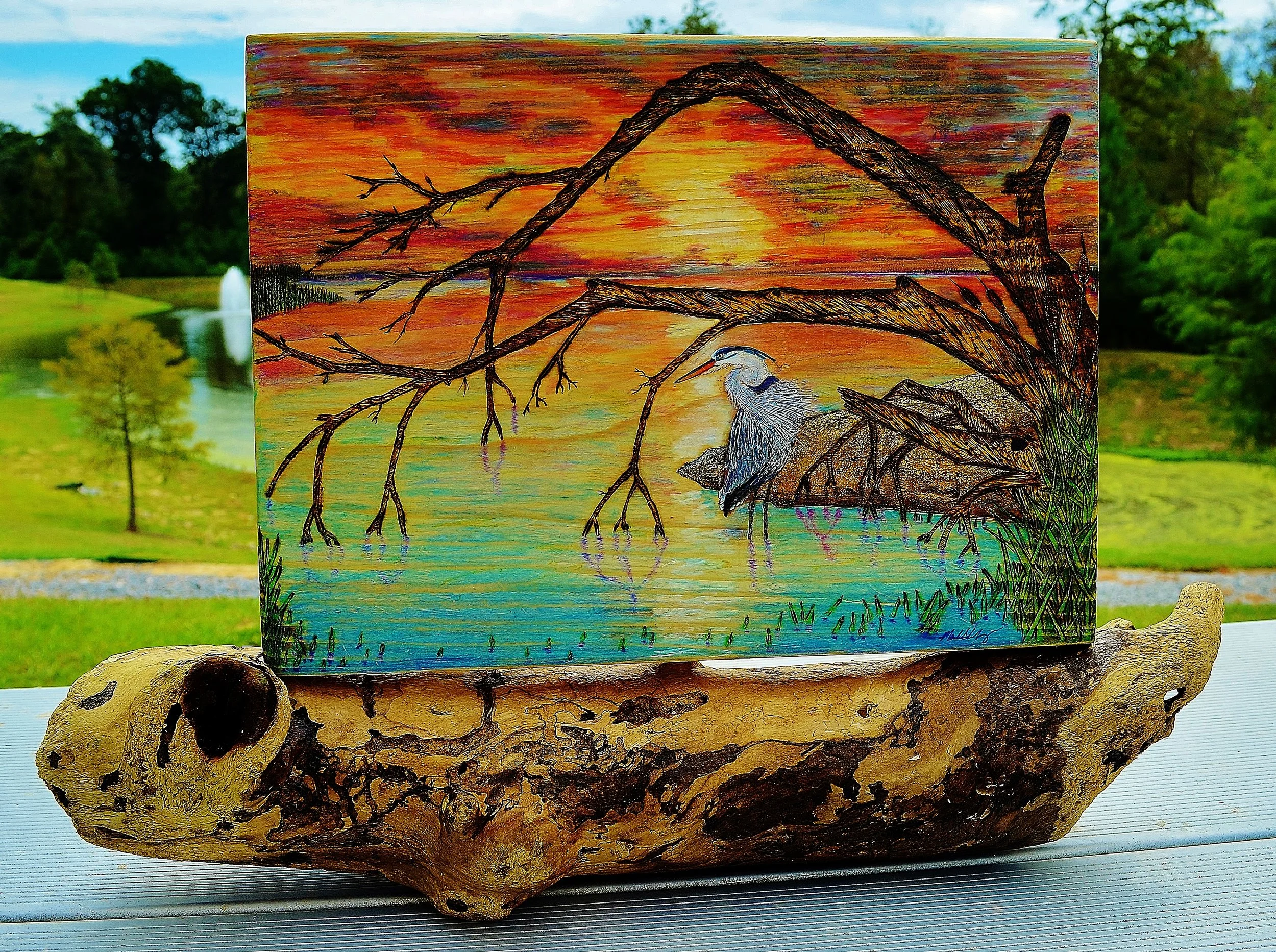 Great Blue Heron on Lake Pontchartrain at Sunset by Art Illusions by Young, Folsom, Louisiana, Hand-burned pyrography artwork on wood depicting a great blue heron perched on driftwood against a vibrant sunset sky over Lake Pontchartrain.