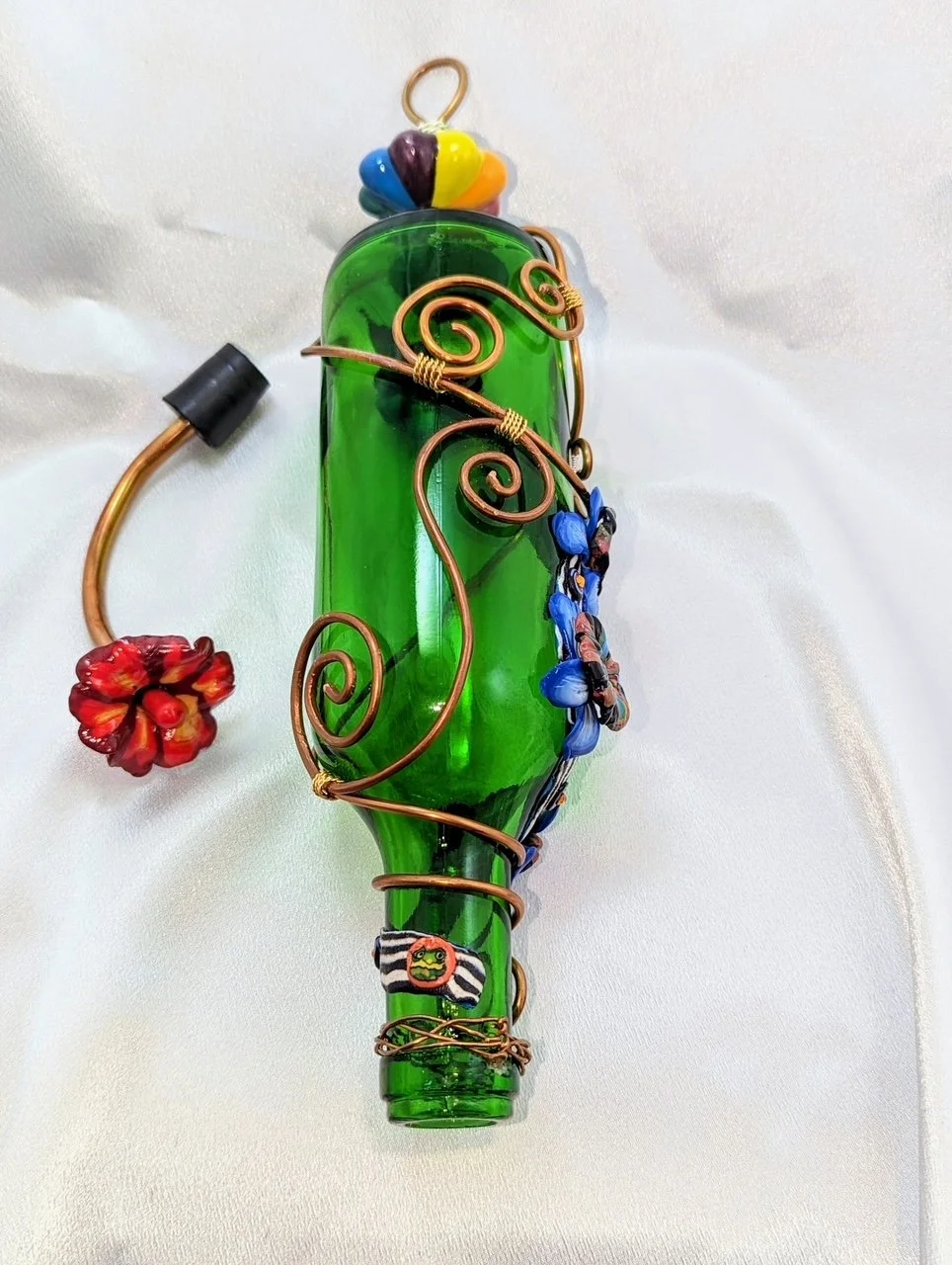 Close-up left side view of the Emerald Garden Bloom Hummingbird Feeder by Art Illusions by Young. The recycled emerald green glass bottle features elegant copper wire wrapping and hand-sculpted blue and multicolored clay flowers, finished with a pill