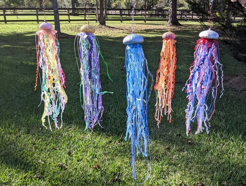 Outdoor garden view of the Patriotic Jellyfish Suncatcher – Stars & Stripes in Painted Motion by Art Illusions by Young, displayed among other pieces in a natural setting. The crisp white sea urchin bell and long, flowing ribbons in vibrant red, whit