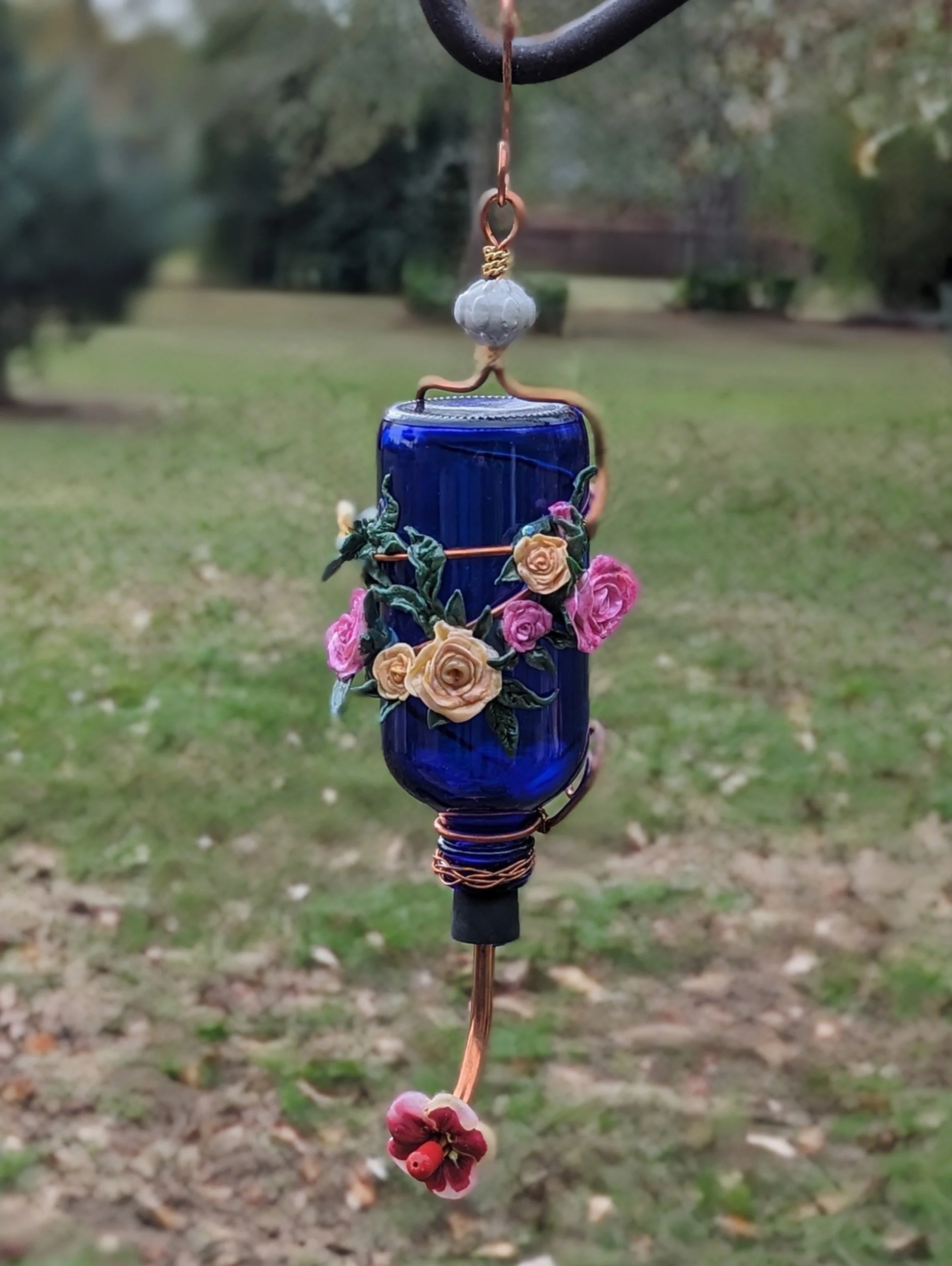 🔴 Sold: 
Outdoor Blue Floral Hummingbird Feeder in Garden Setting by Art Illusions by Young, Folsom, Louisiana. Shown in its natural garden environment, this cobalt blue glass hummingbird feeder displays lush hand-sculpted polymer clay roses and blo
