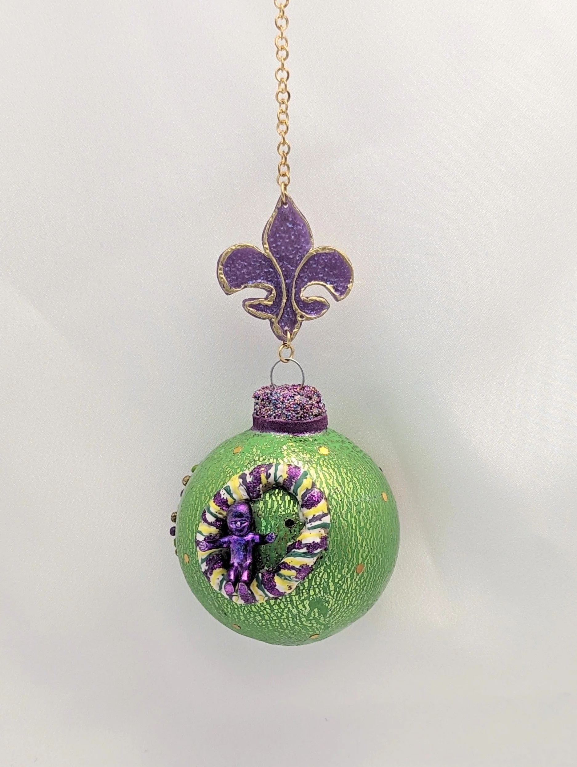 🔴 Sold: 
Green Christmas Ornament by Art Illusions by Young. A festive green spherical ornament with sparkling accents and a decorative Mardi Gras King Cake and baby. Handcrafted in polymer clay, it brings bright holiday color and elegant detail to 