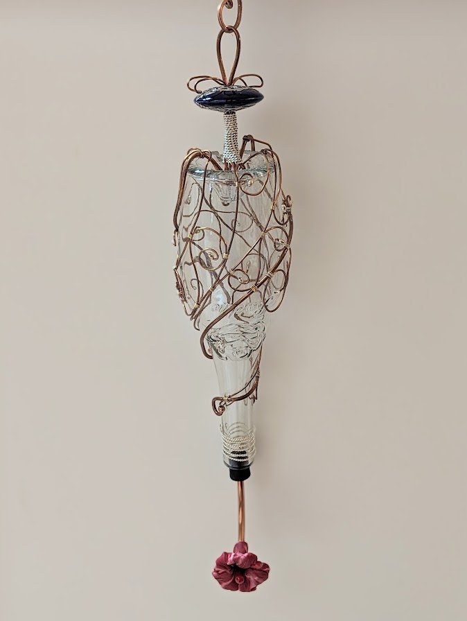 Hanging view of the Copper Spiral Grace Hummingbird Feeder by Art Illusions by Young. The recycled clear glass bottle features sophisticated copper wire wrapping formed into artistic spirals and loops, finished with a secure decorative cobalt ceramic