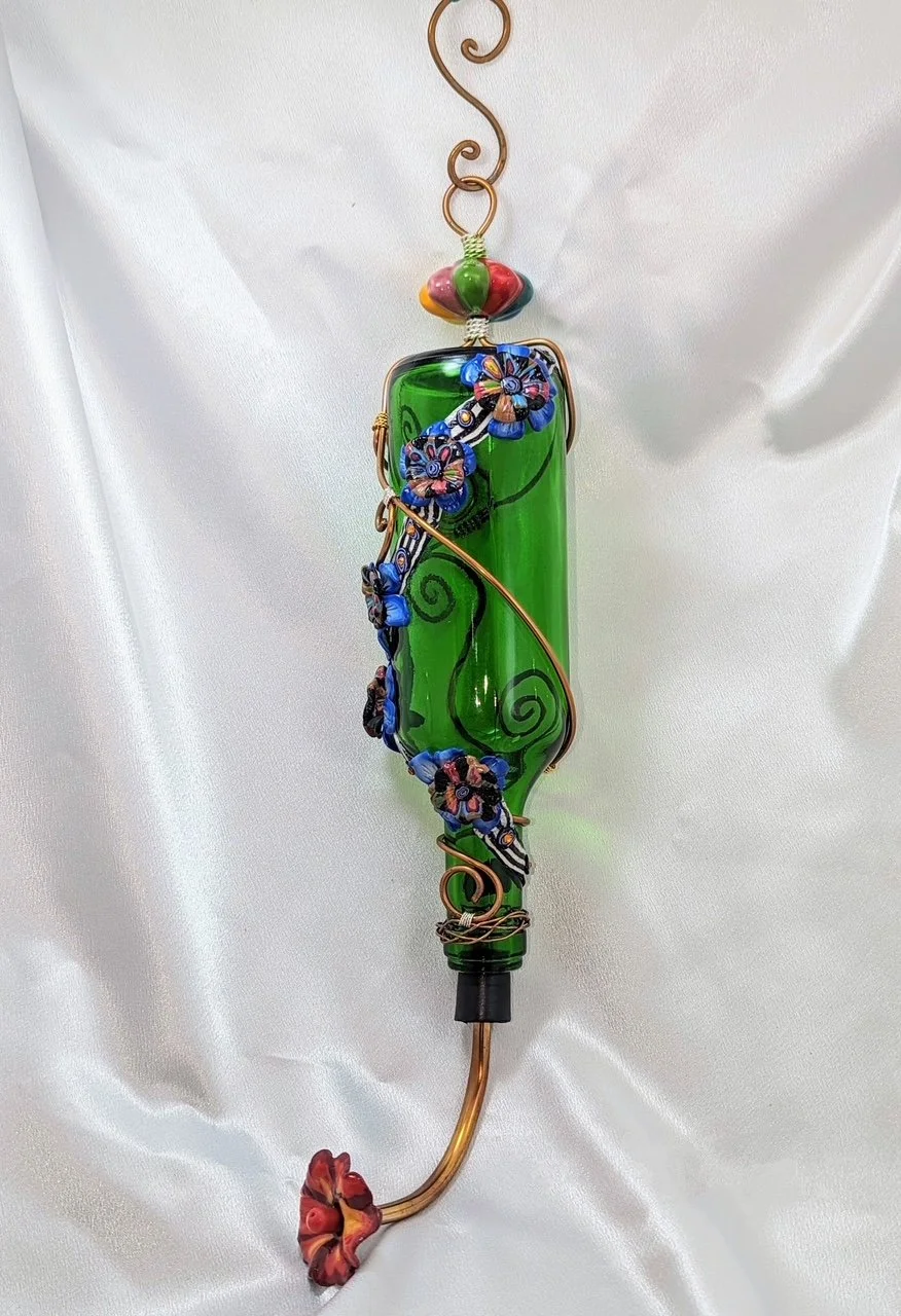 Green glass hummingbird feeder by Art Illusions by Young adorned with blue flower embellishments and copper wire accents, shown against a white background.
