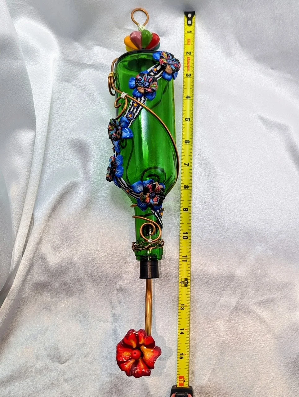 Green glass hummingbird feeder by Art Illusions by Young with colorful floral and wire decorations, shown with a yellow ruler for scale against a white background.