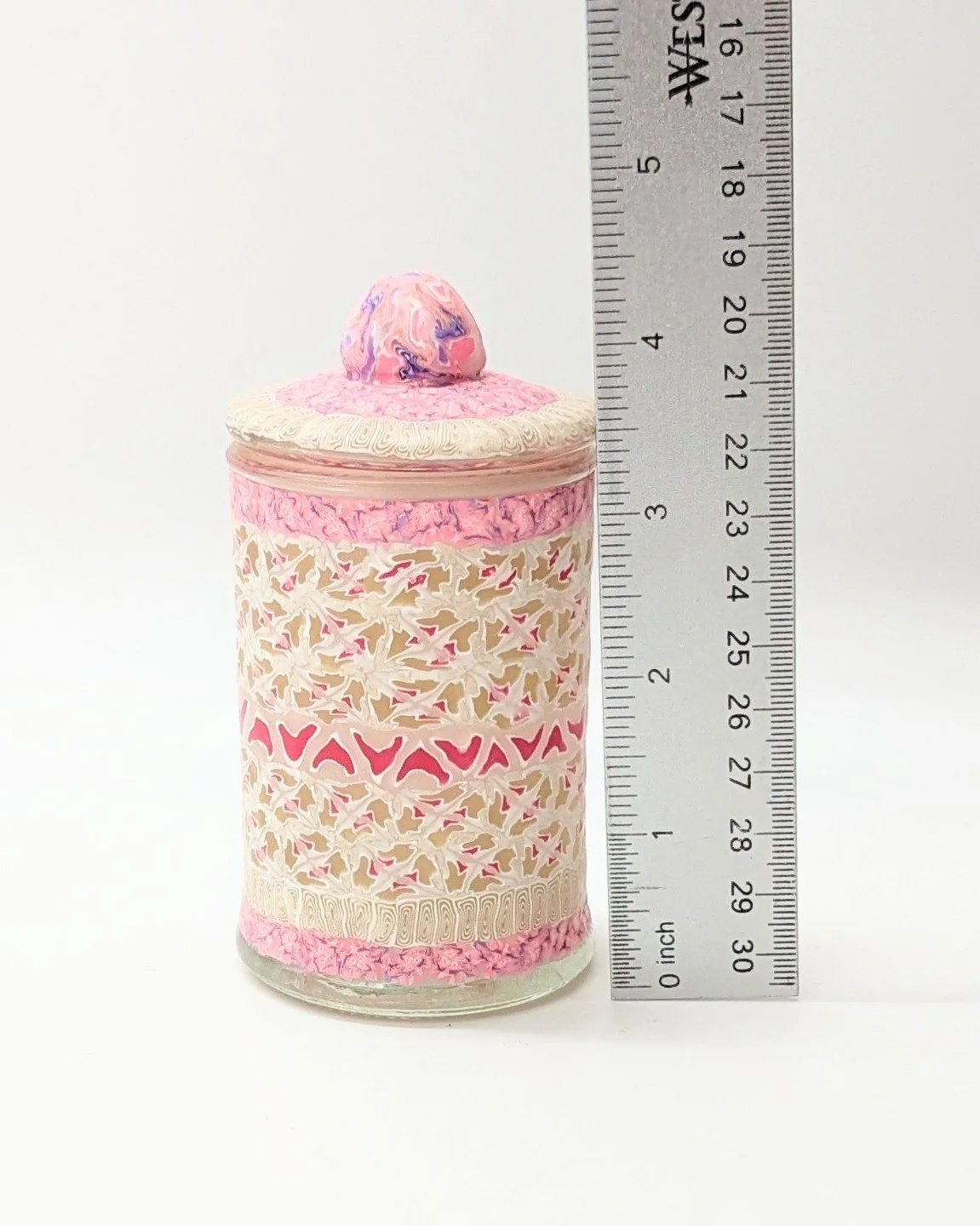 Side profile of the Floral Lace Trinket Jar by Art Illusions by Young, shown with a ruler for scale. The intricate hand-sculpted lace-like patterns in soft cream, accented with pink and gold, wraps seamlessly around the cylindrical form. The domed li