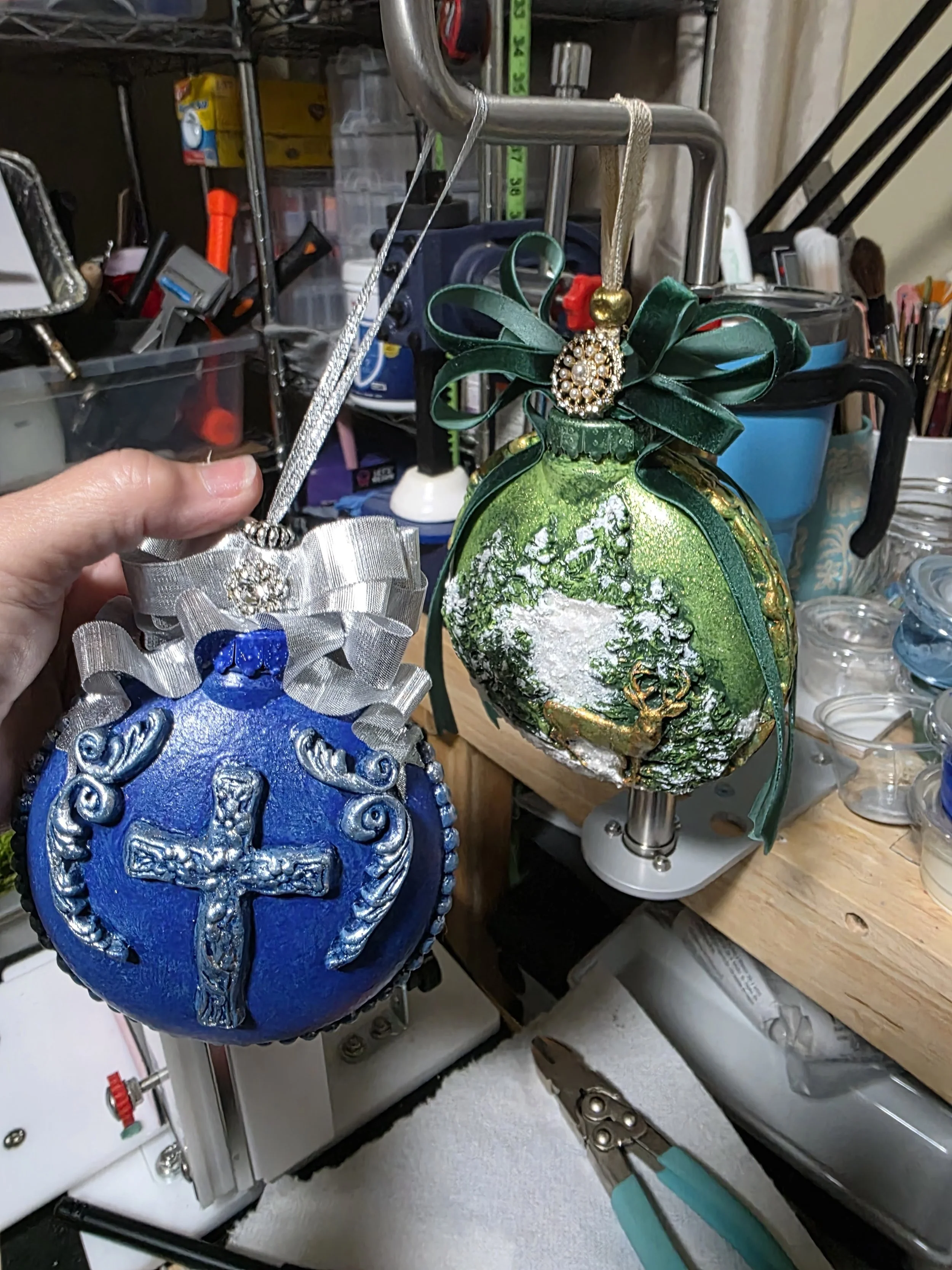 Studio process view of the Woodland Majesty Ornament by Art Illusions by Young. The artist holds two different in process pieces alongside tools and materials. This behind-the-scenes image highlights the meticulous texturing, painting, and assembly.

