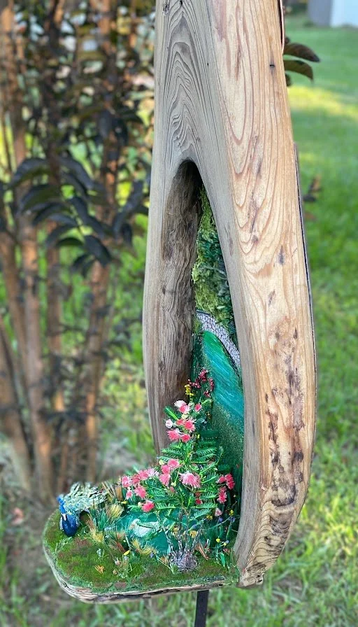 Interior close-up of the miniature park scene inside the Enchanted City Park Serenity diorama, showing the reflective pond, arched bridge, and abundant pink mimosa flowers. Hand-sculpted details by Art Illusions by Young.