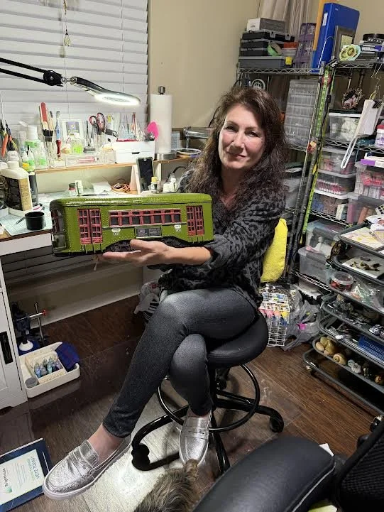 Sandra Young of Art Illusions holding the hand-crafted New Orleans St Charles Avenue streetcar diorama in Folsom, Louisiana studio. Olive green streetcar with crimson windows, glowing LED lights, functional tissue box design on tracks with flowers.