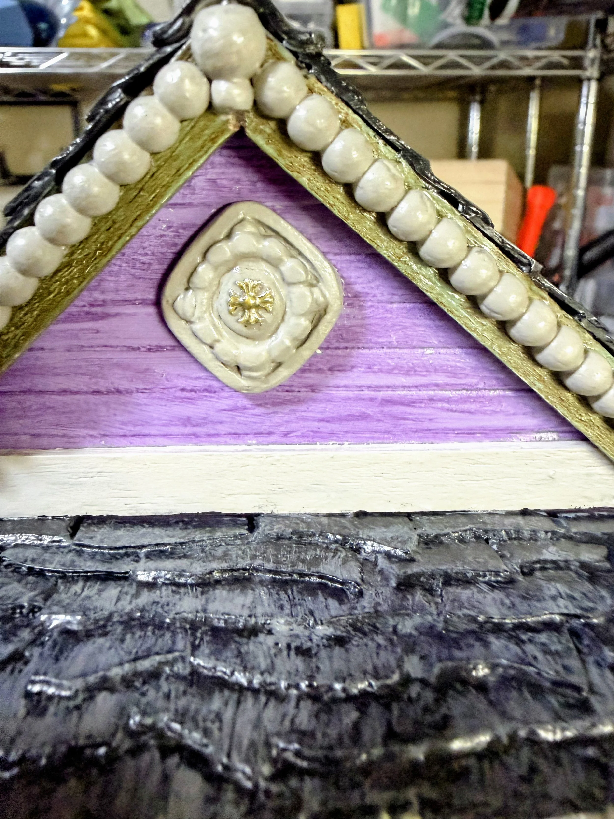 Extreme close-up of the Purple Reign Shotgun House roof peak by Art Illusions by Young. Features the golden cross, decorative diagonal carving, slate shingles, and ornate gingerbread trim. Hand-sculpted polymer clay and wood details on this functiona