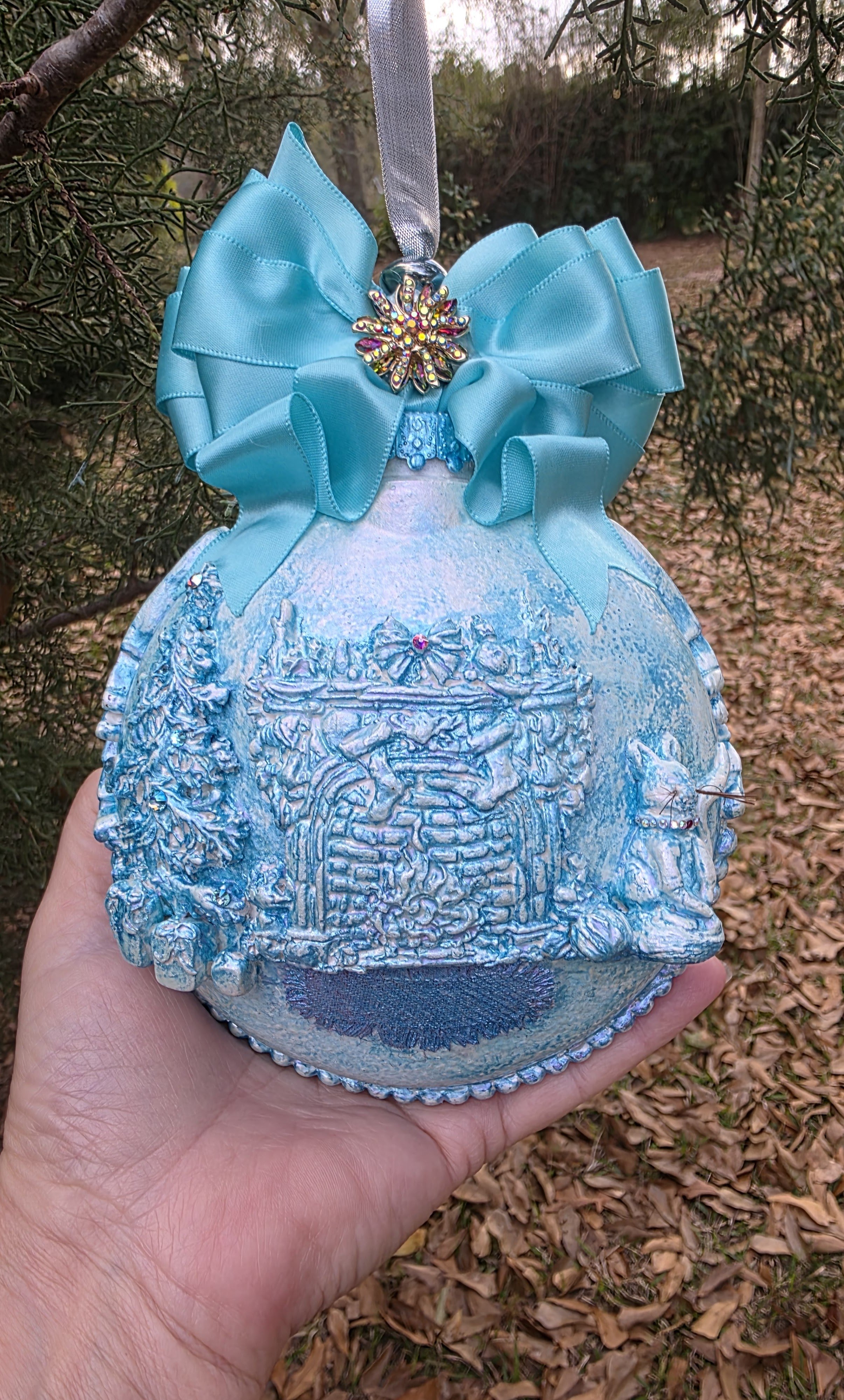 Artist's hand holding the Cozy Fireplace with Cat Ornament by Art Illusions by Young for scale. The intricate and sparkling texture in soft light turquoise blue tones covers the spherical form, while a large aqua satin ribbon bow with jeweled accent 