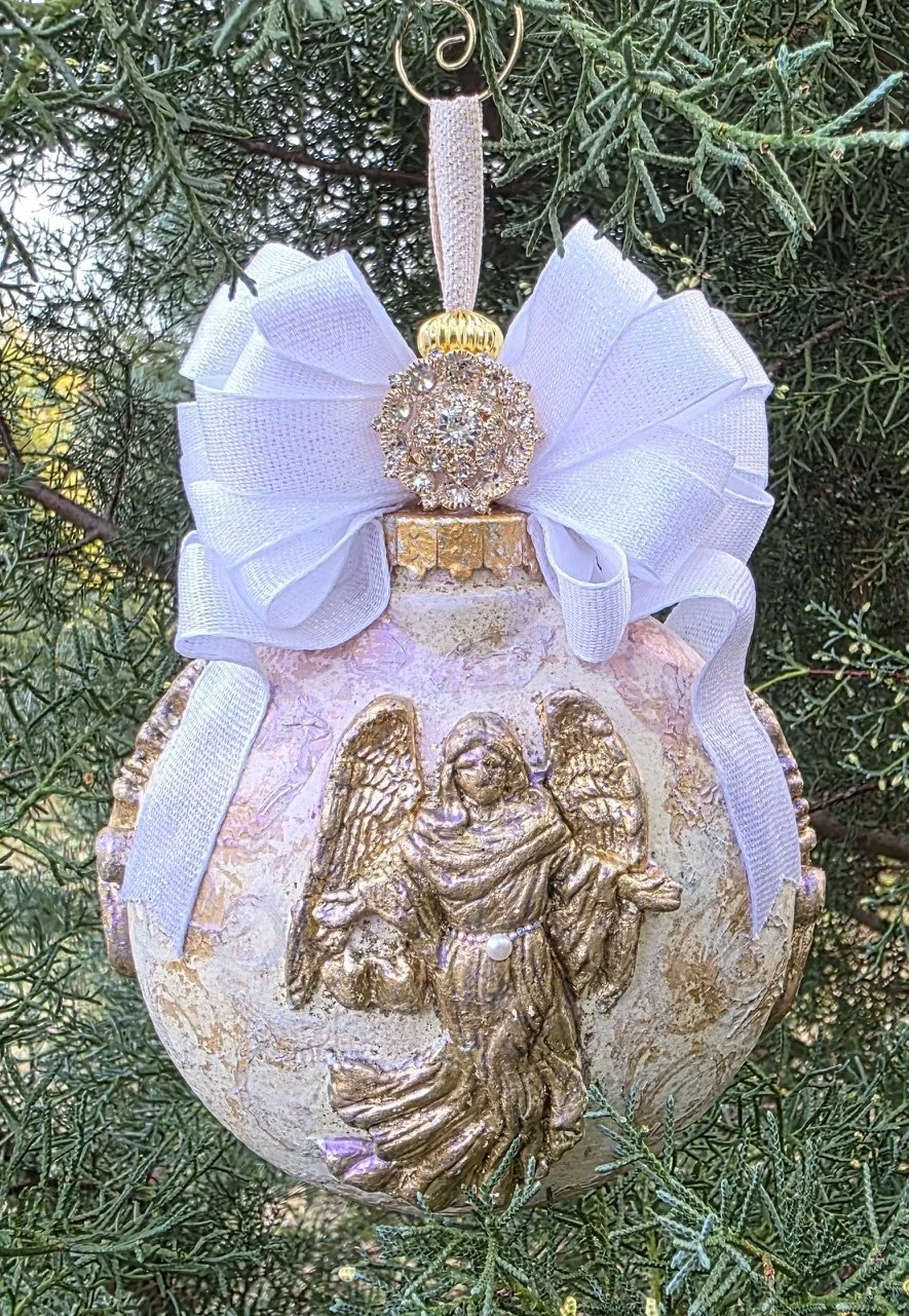 A hand-sculpted and molded Guardian Angel Ornament by Art Illusions by Young hangs gracefully among lush evergreen branches. The detailed angel figure, rendered in soft iridescent pearl, blush, and golden tones, features outstretched wings and flowin