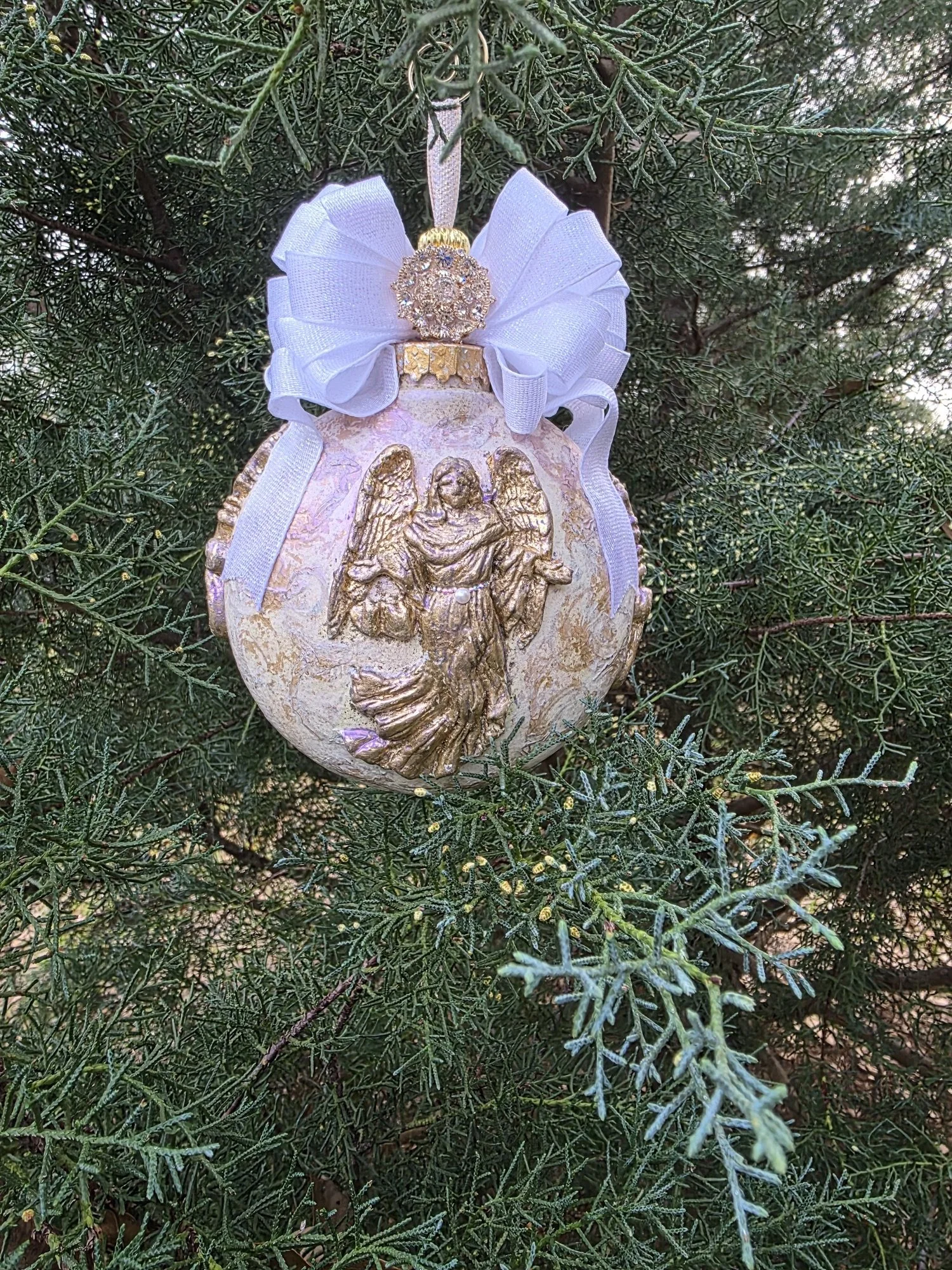 

Outdoor view of the Golden Guardian Angel Ornament by Art Illusions by Young. 
The angel stands as a serene guardian, surrounded by subtle scrollwork and metallic accents that enhance the ethereal quality of the surface. Crowned with a 
 generous w