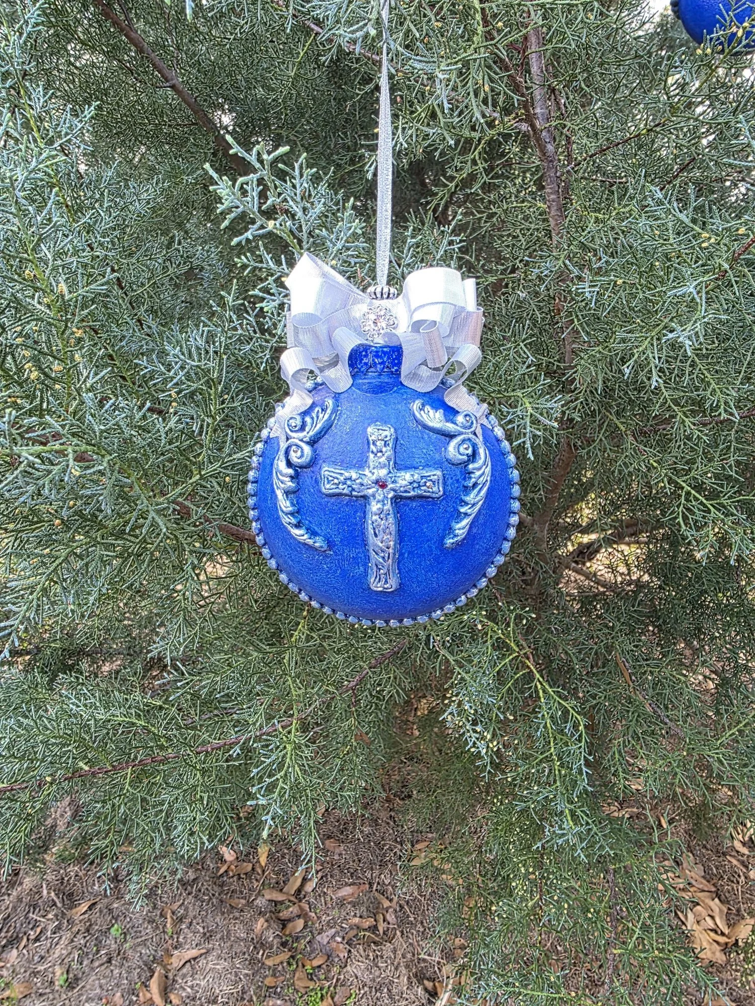 Hanging view of the Azure Blue Cross Ornament by Art Illusions by Young on a tree. The compact beautiful blue orb showcases the intricate silver cross and raised flourishes, crowned by a sparkling silver ribbon bow. This versatile ornament radiates r
