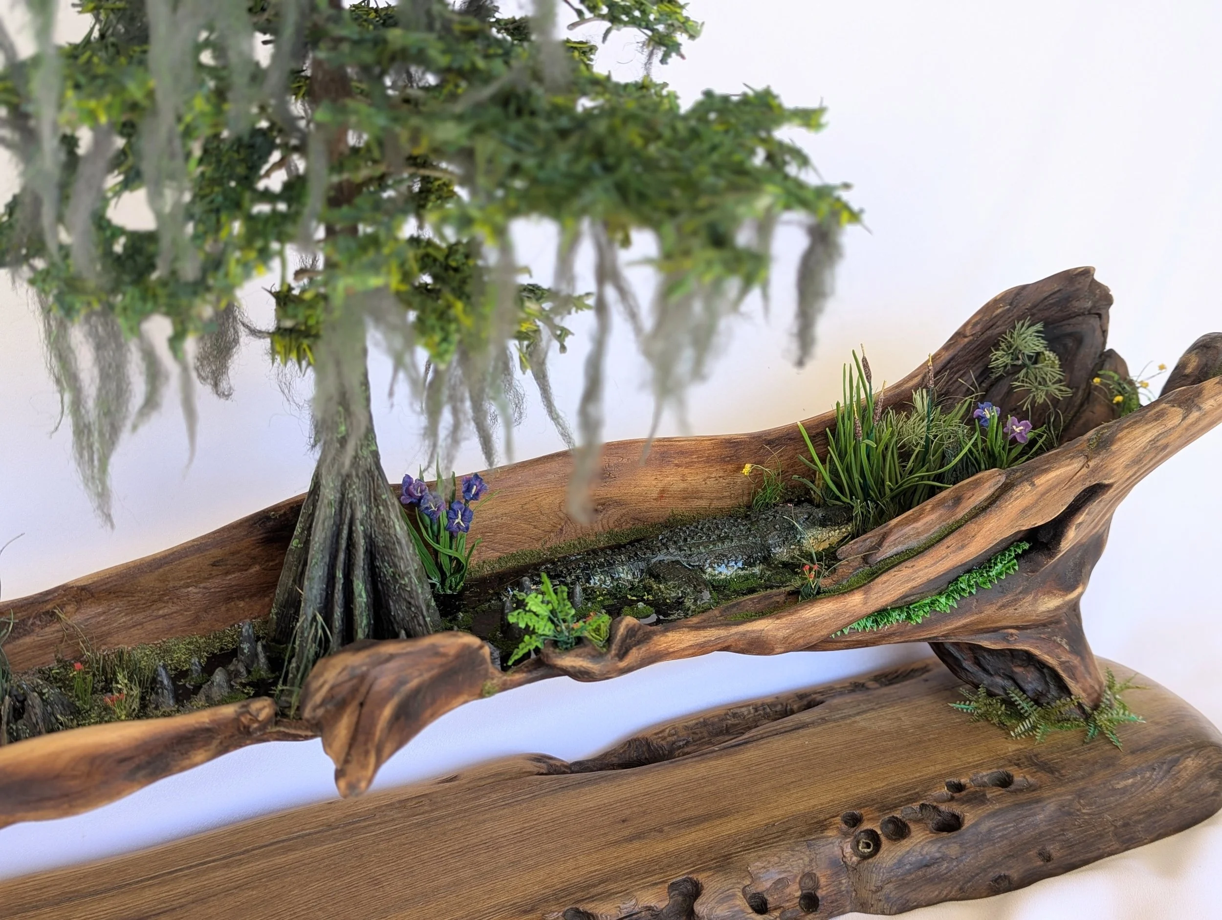 Driftwood base with vibrant green moss and emerging plants in a micro swamp diorama. Detailed handcrafted work by Art Illusions by Young.