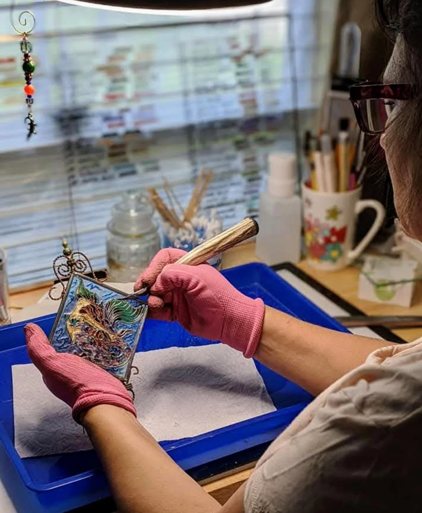 Sandra Young wearing pink gloves carefully working on a colorful mermaid design in a decorative piece at the Art Illusions by Young studio.
