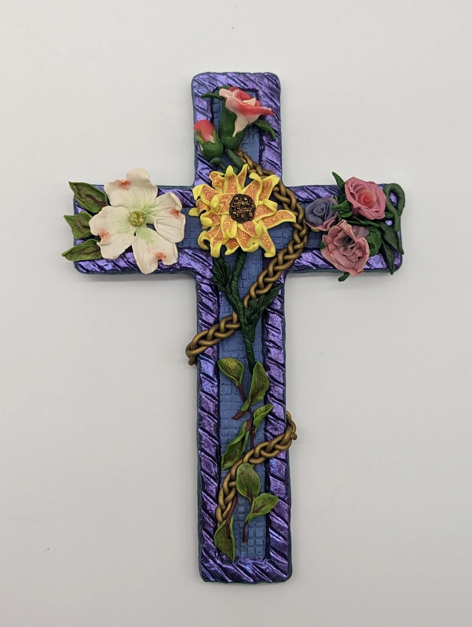 🔴 Sold: 
Floral Cross by Art Illusions by Young, Folsom, Louisiana. This striking sculptural cross features a rich purple textured background with a prominent golden braid running vertically and horizontally. Hand-sculpted polymer clay flowers adorn