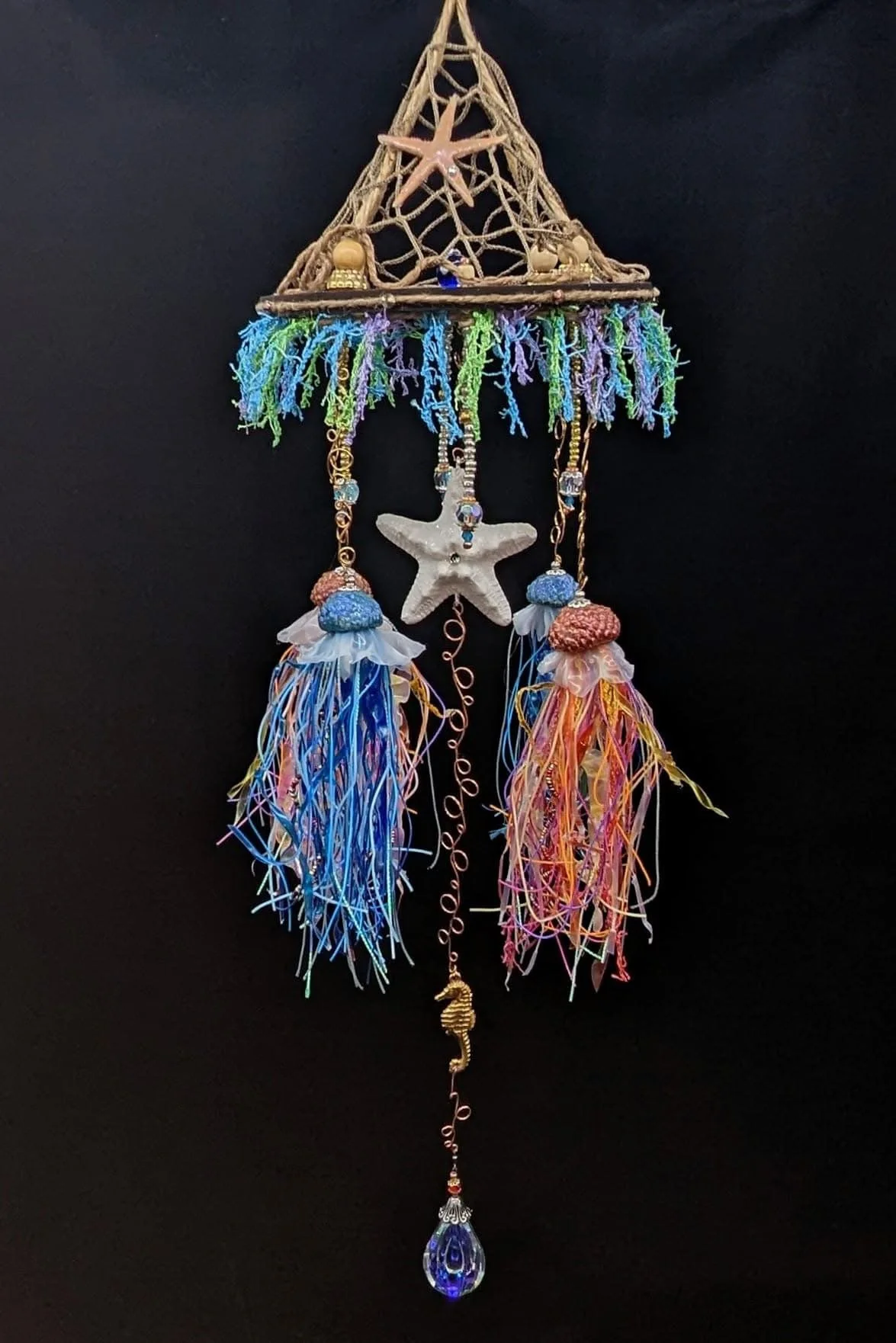 Angled outdoor view of the Jellyfish Mobiles – Chihuly-Inspired Light Catcher by Art Illusions by Young. The scrollwork wooden hanger and decorative netted canopy suspend four handcrafted jellyfish with flowing tentacles, while resin-sealed starfish,