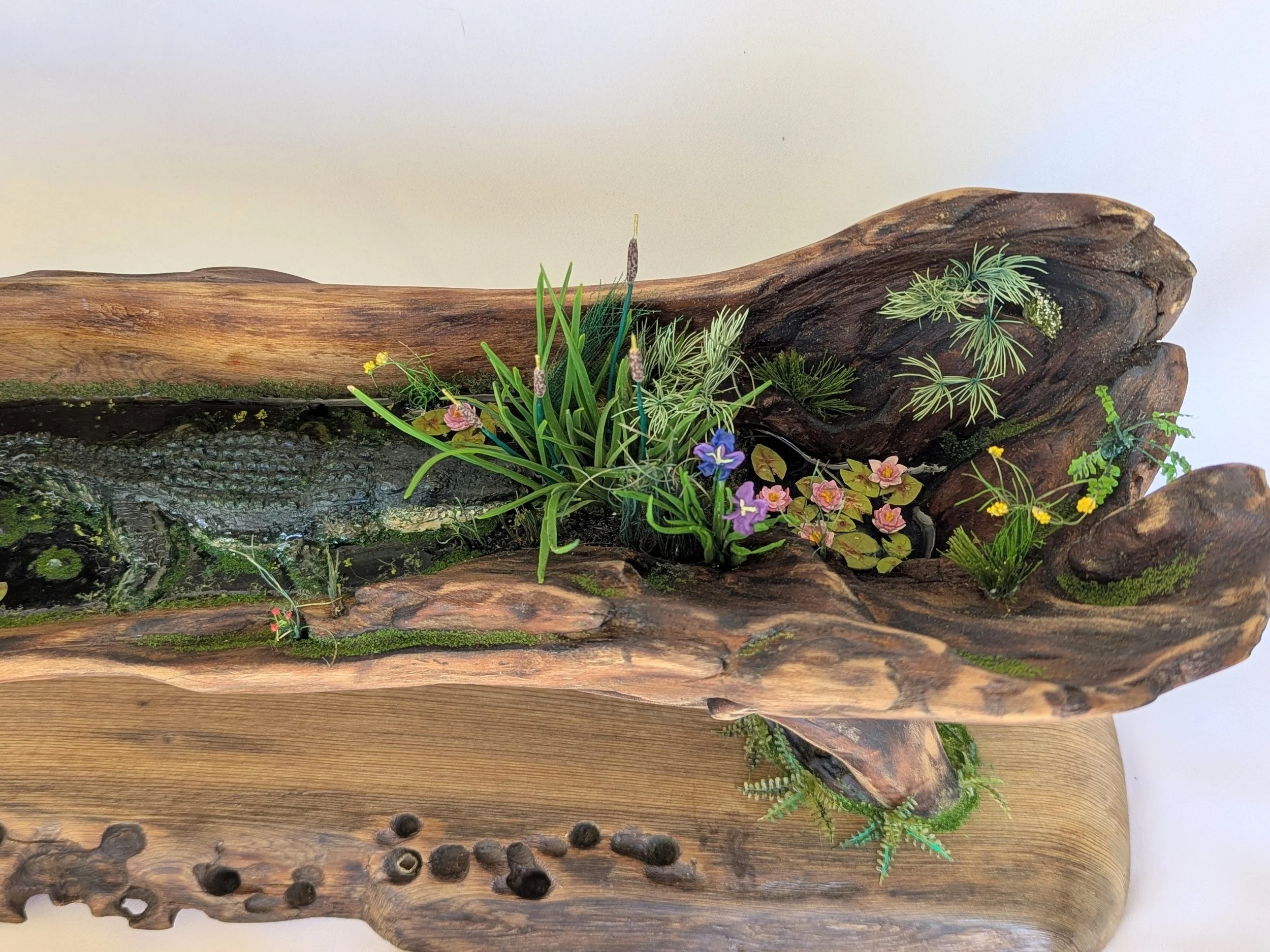 Handcrafted micro swamp diorama by Art Illusions by Young featuring a small sculpted alligator resting among green plants, cattails, blue flowers, and reflective black epoxy water on a natural driftwood base. Realistic Louisiana bayou wetland scene f
