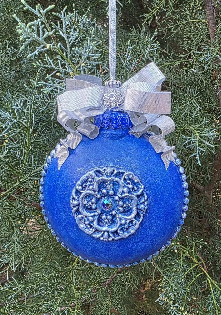 Rear view of the Azure Blue Cross Ornament by Art Illusions by Young. The ornate silver cross with delicate filigree and scrollwork stands in elegant contrast to the rich blue base with its subtle metallic sheen. A dramatic silver double-sided ribbon