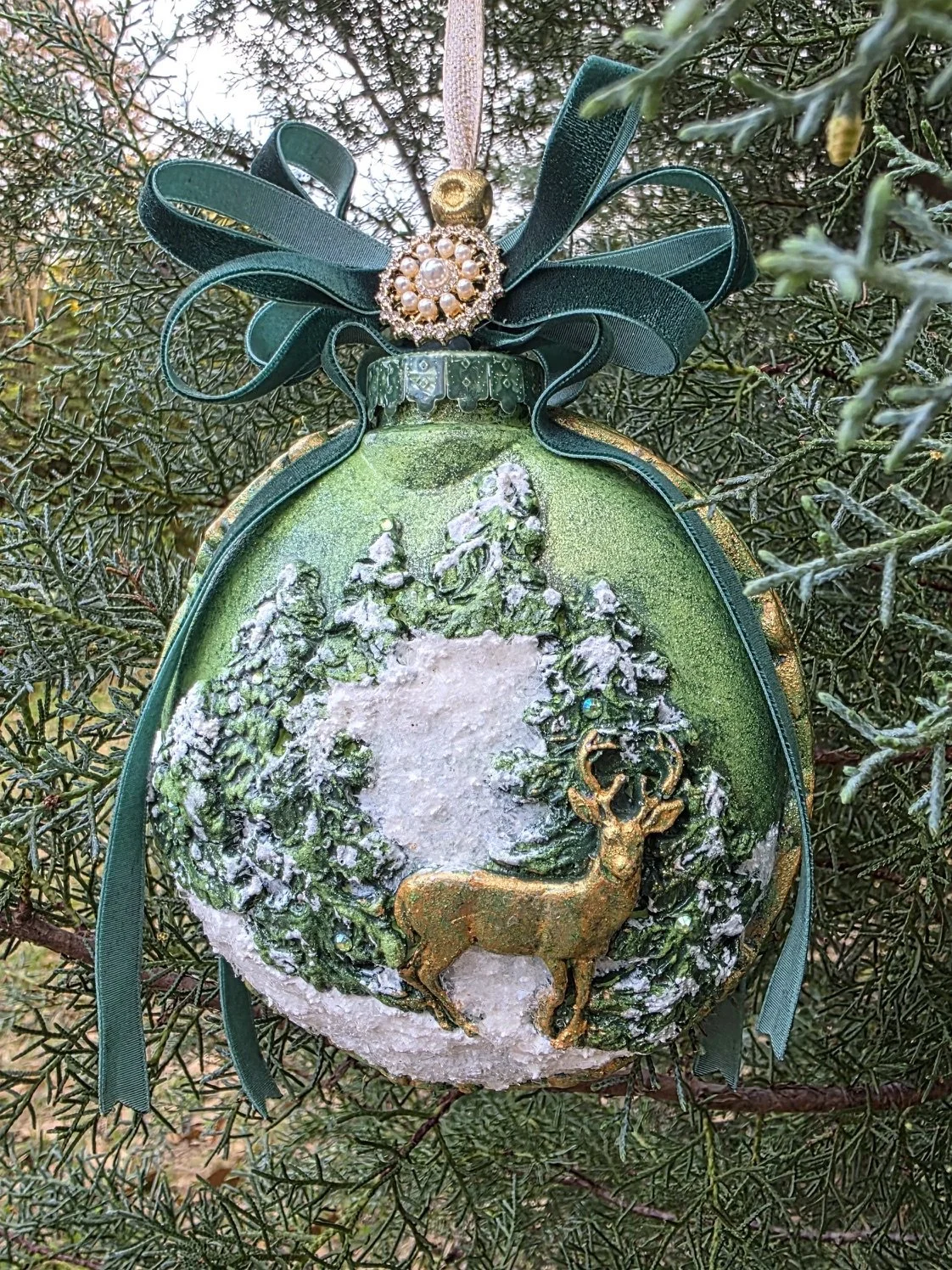 Hanging view of the Woodland Majesty Ornament by Art Illusions by Young on a tree. The spherical form showcases the detailed stag and winter woodland scene in rich metallic green tones, crowned by a generous dark green ribbon bow. This hand-sculpted 