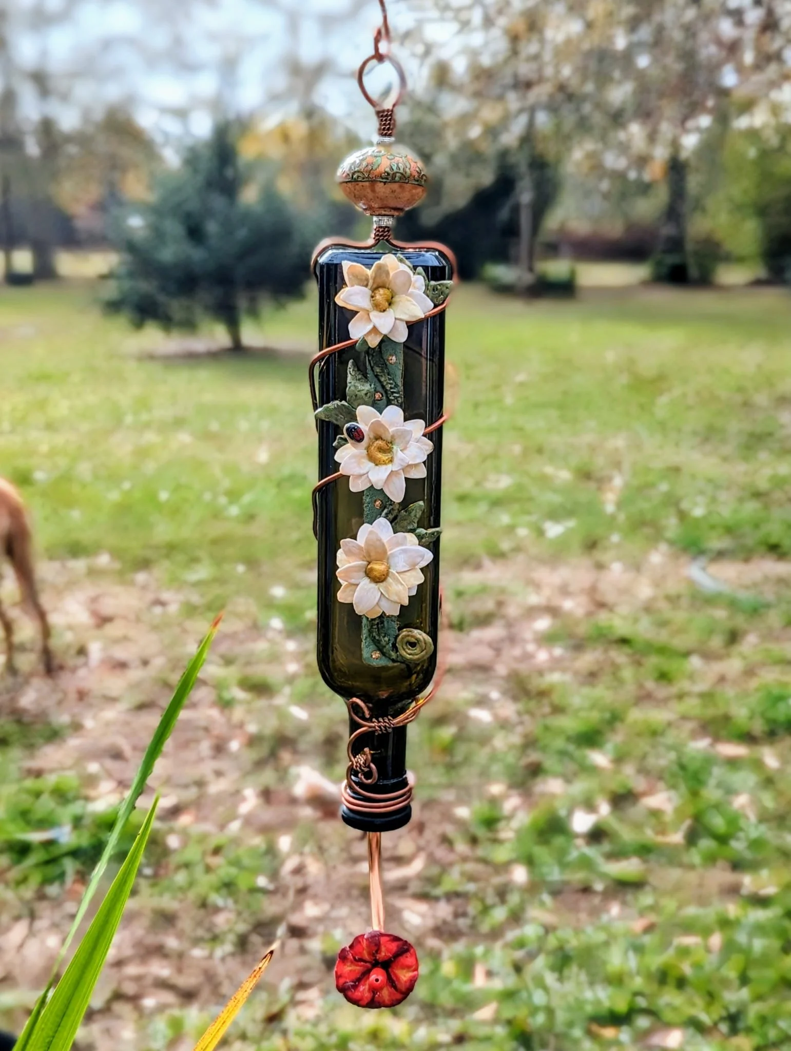 Handmade decorative hummingbird feeder with white flowers and green leaves by Art Illusions by Young. Suspended from a copper wire with a red flower at the bottom, displayed against a grassy outdoor background.