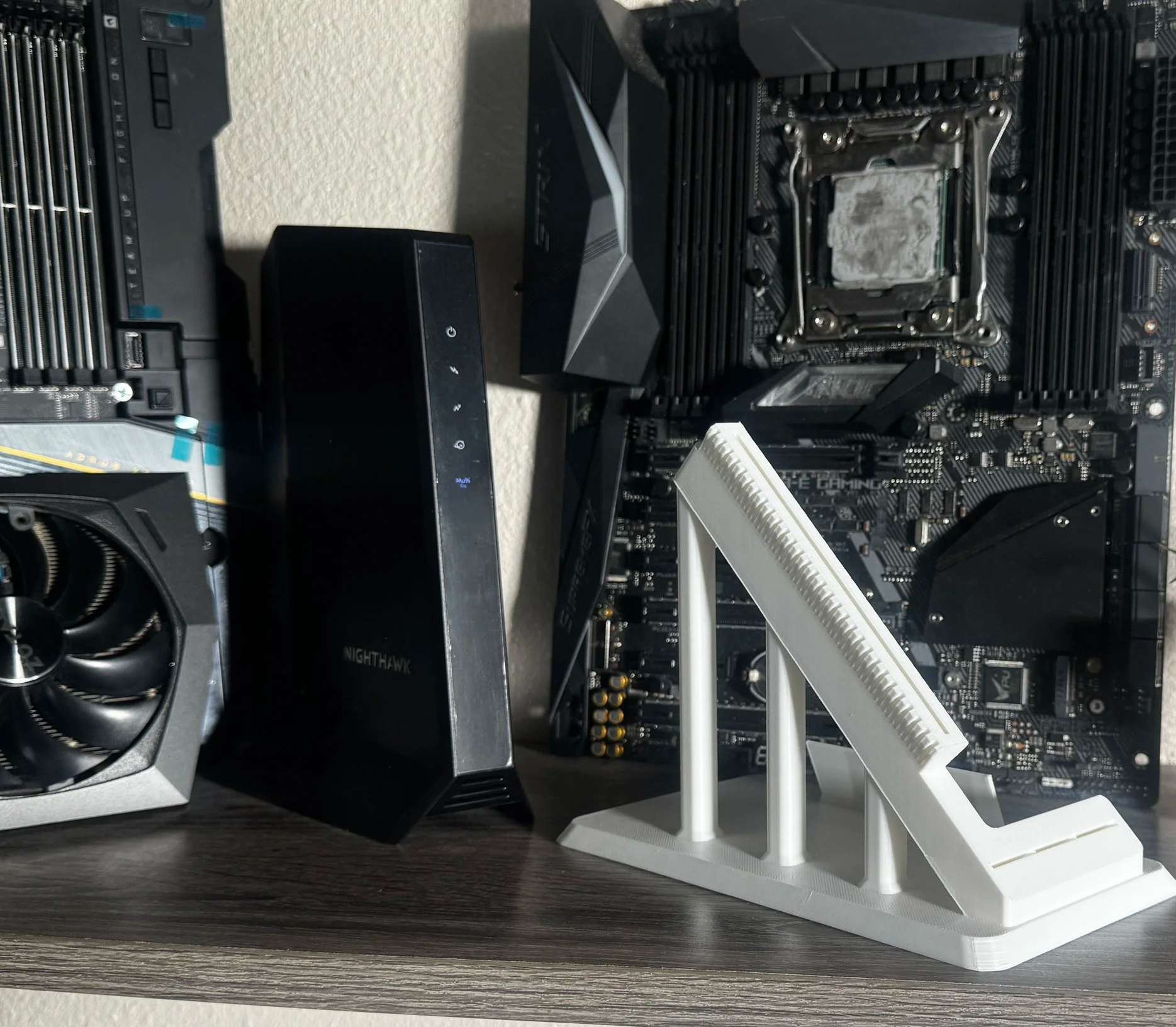 Close-up of a computer motherboard with a CPU socket, RAM slots, and other components. An anti-static stand is in the foreground, and a graphics card, a wireless router, and a black power supply are also visible.