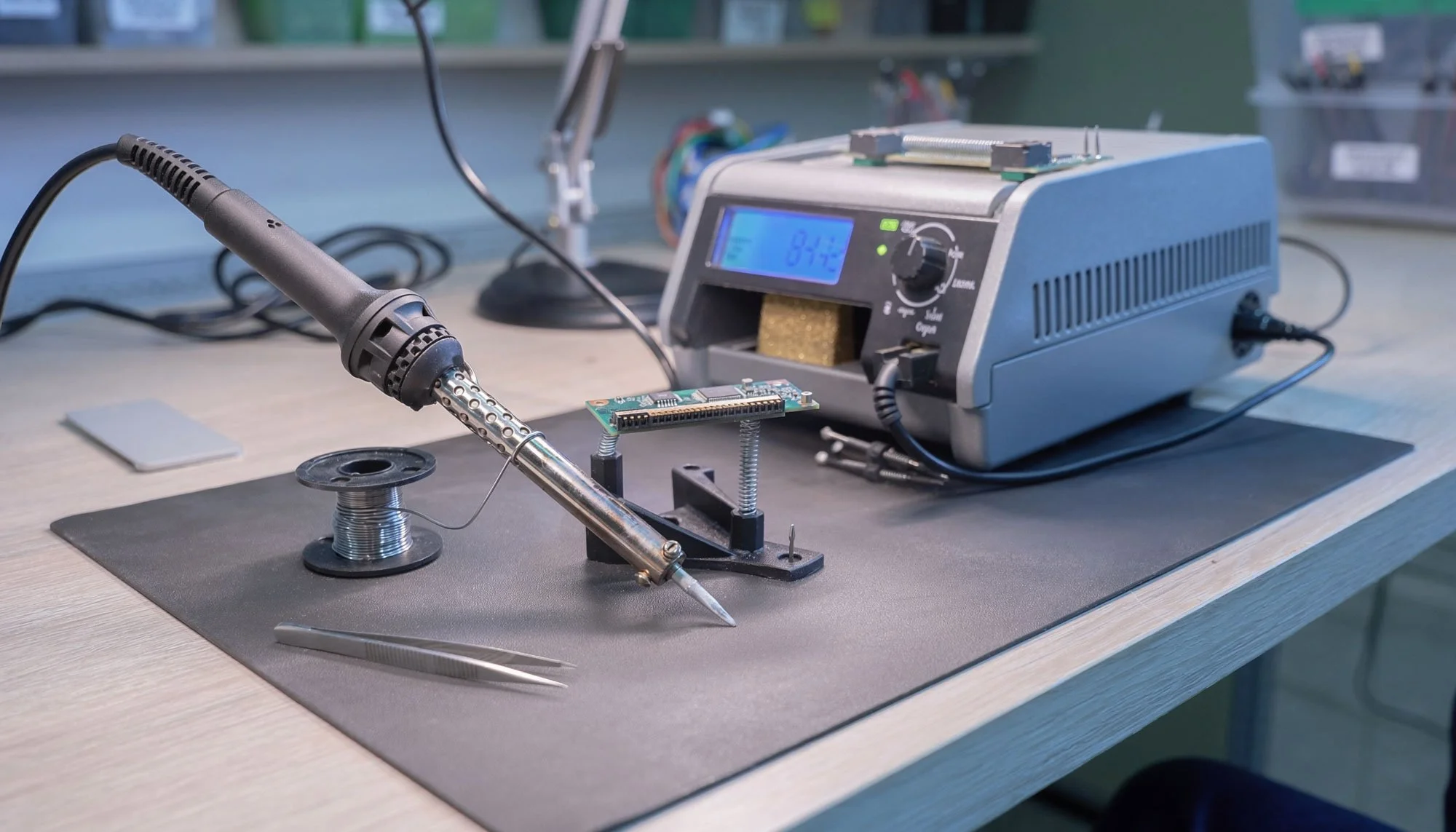 Soldering station with a soldering iron, spool of solder, and a circuit board on a workbench.