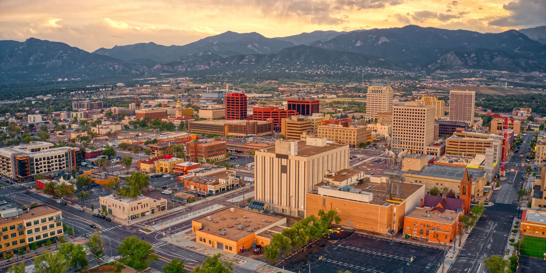 A cityscape at sunset with tall buildings, mountains in the background, and mostly empty streets in the foreground.