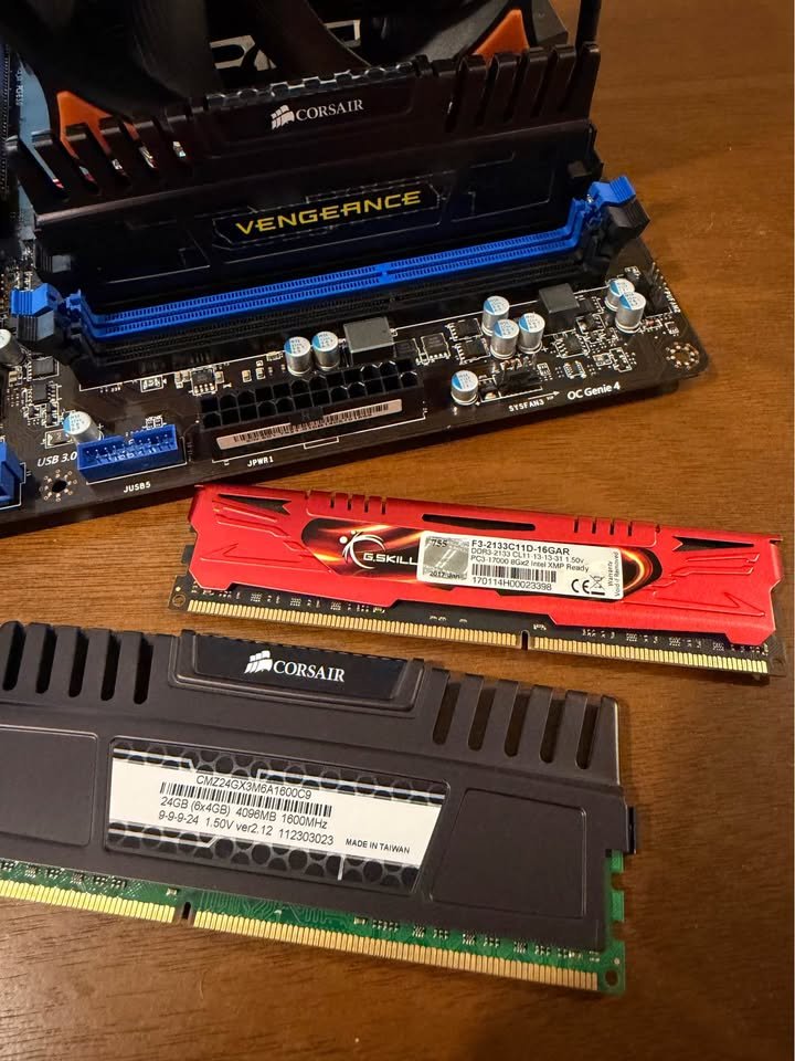Photo of computer RAM modules and a motherboard. The modules include Corsair Vengeance and G.Skill RAM, with each labeled with specifications. The motherboard has a Corsair branding and a visible heatsink.