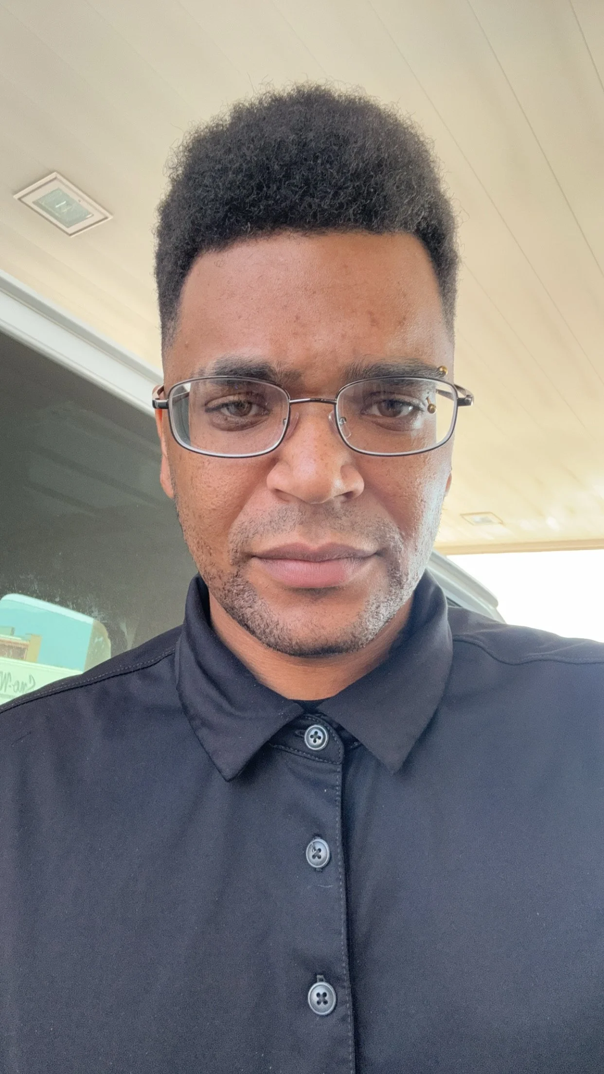 A man with glasses wearing a black collared shirt, standing outdoors in front of a building with a wooden roof, looking at the camera.