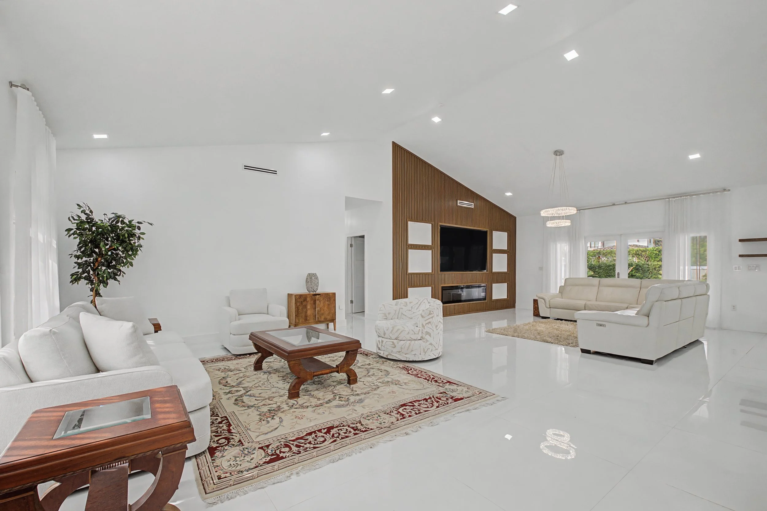 Bright spacious living room with white walls, white tile floor, and large windows with sheer white curtains. Contains white sofas, a wooden coffee table, a patterned armchair, a beige area rug, and a built-in wooden entertainment center with a flat-s
