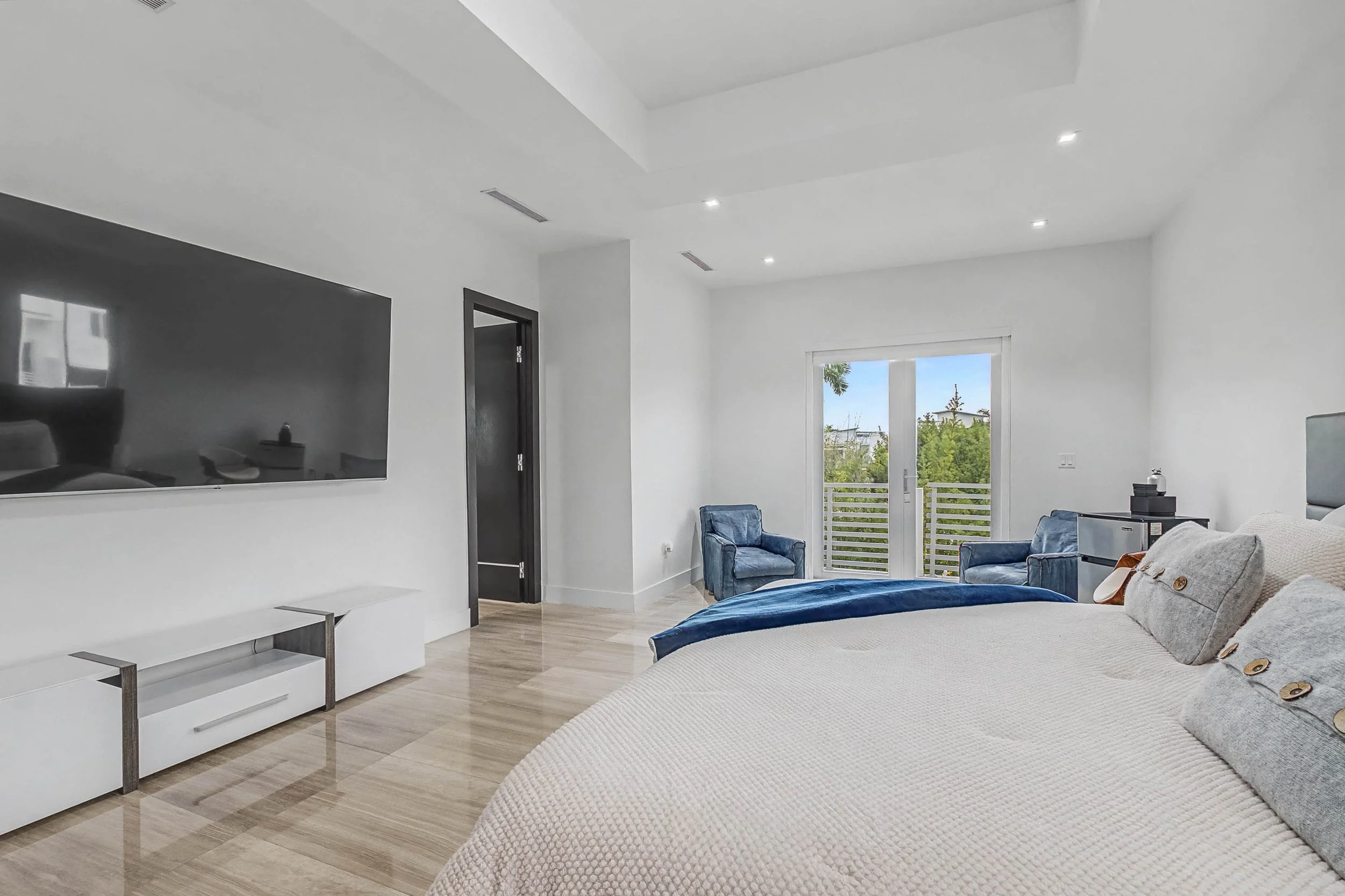 Modern bedroom with white walls, a large flat-screen TV mounted on the wall, a bed with beige bedding and gray pillows, two blue armchairs by a balcony door, and light-colored hardwood floors.