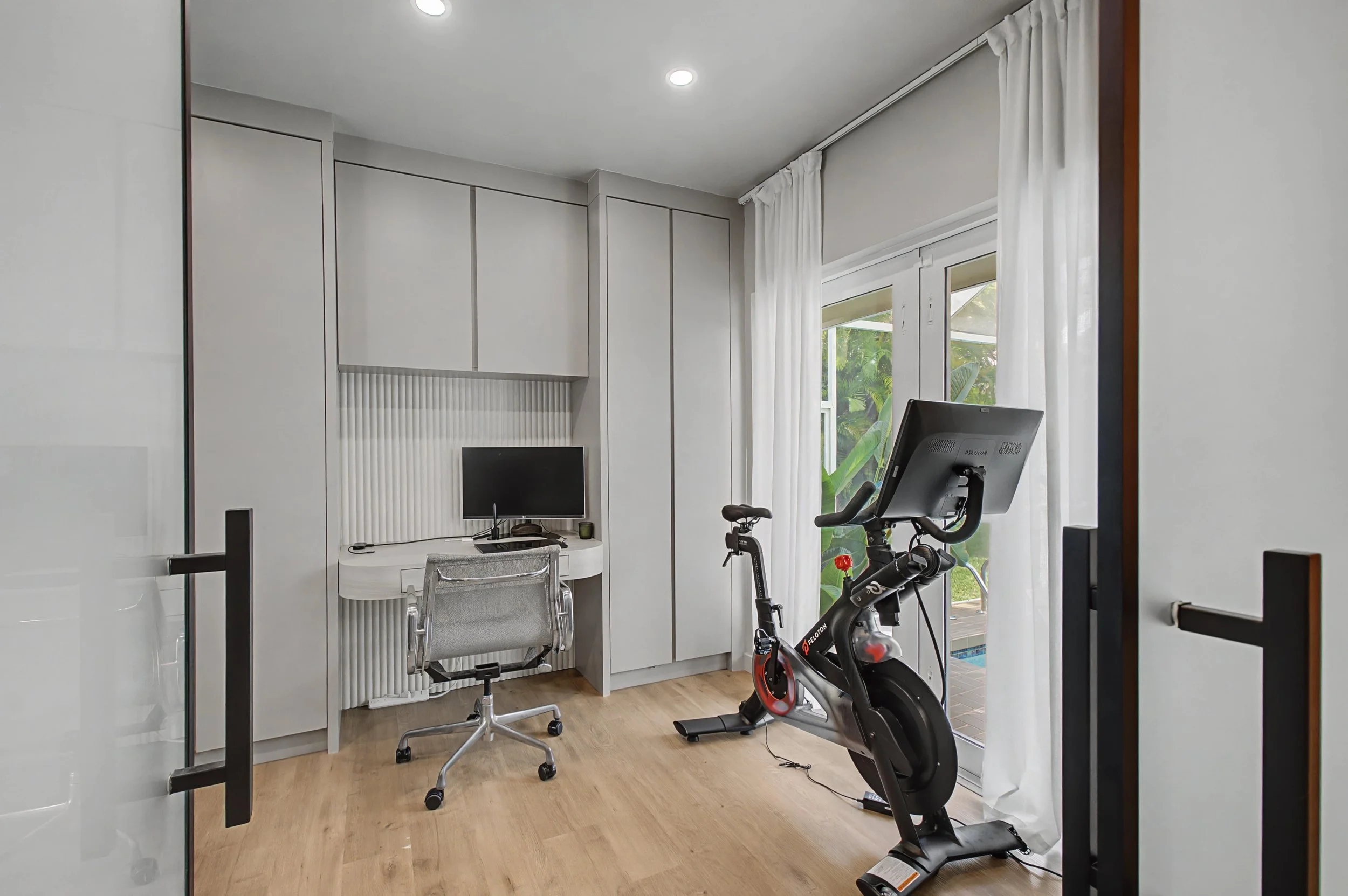 Home office with a desk, chair, and computer monitor next to a stationary exercise bike near glass doors with white curtains, overlooking a tropical garden.