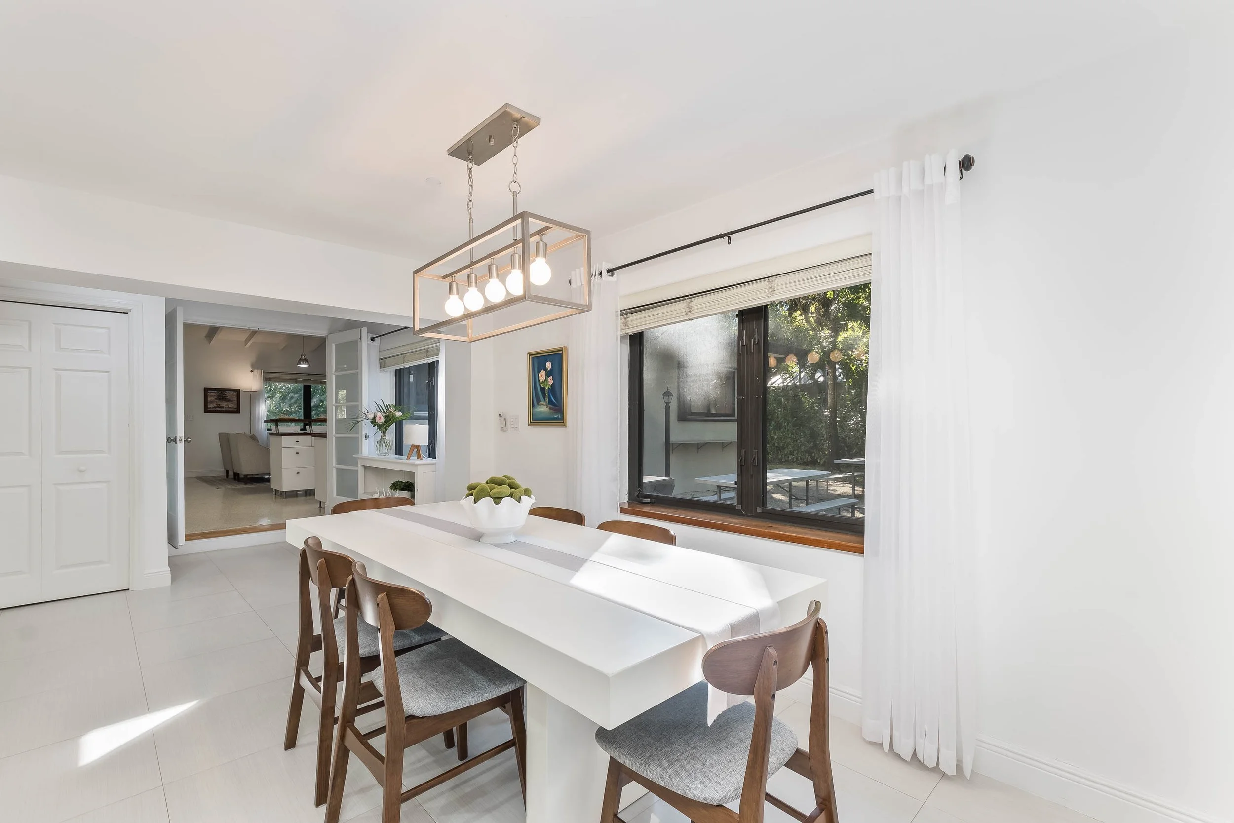 Bright dining room with white walls, a large white table, six wooden chairs with gray cushions, a modern light fixture, a window with white curtains, and a view of the outdoor backyard.