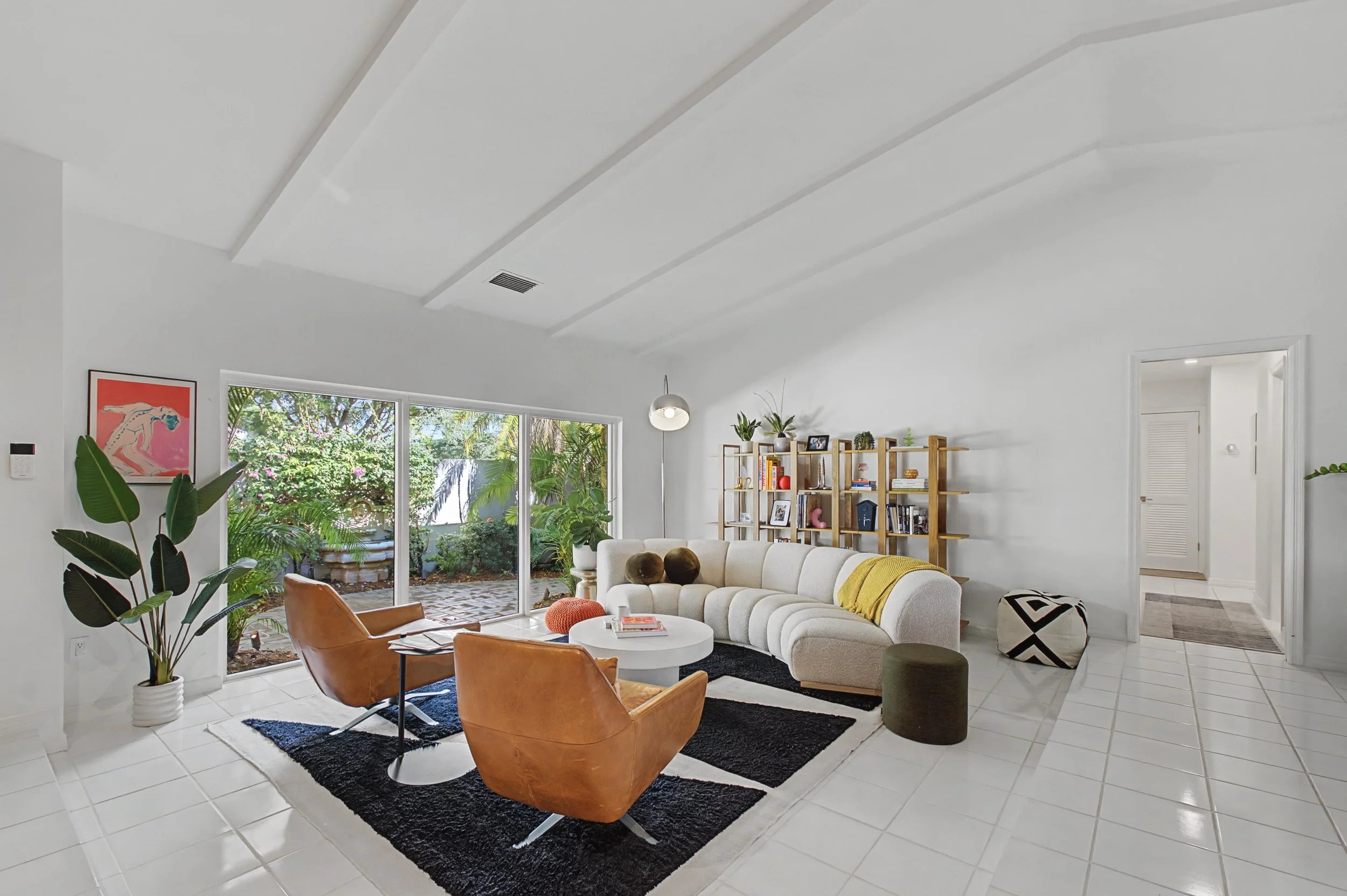 Bright living room with white walls, tile floor, large sliding glass doors leading outside, white curved sofa, two brown leather chairs, white coffee table, black area rug, wooden bookshelf with decorations, potted plant, artwork on wall, and an open