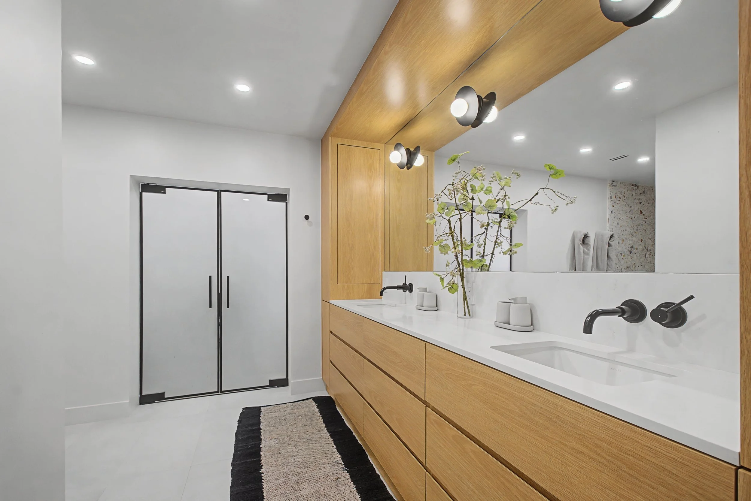 Modern bathroom with wooden vanity, white countertop, black fixtures, large mirror, potted plant, patterned rug, and frosted glass double door.