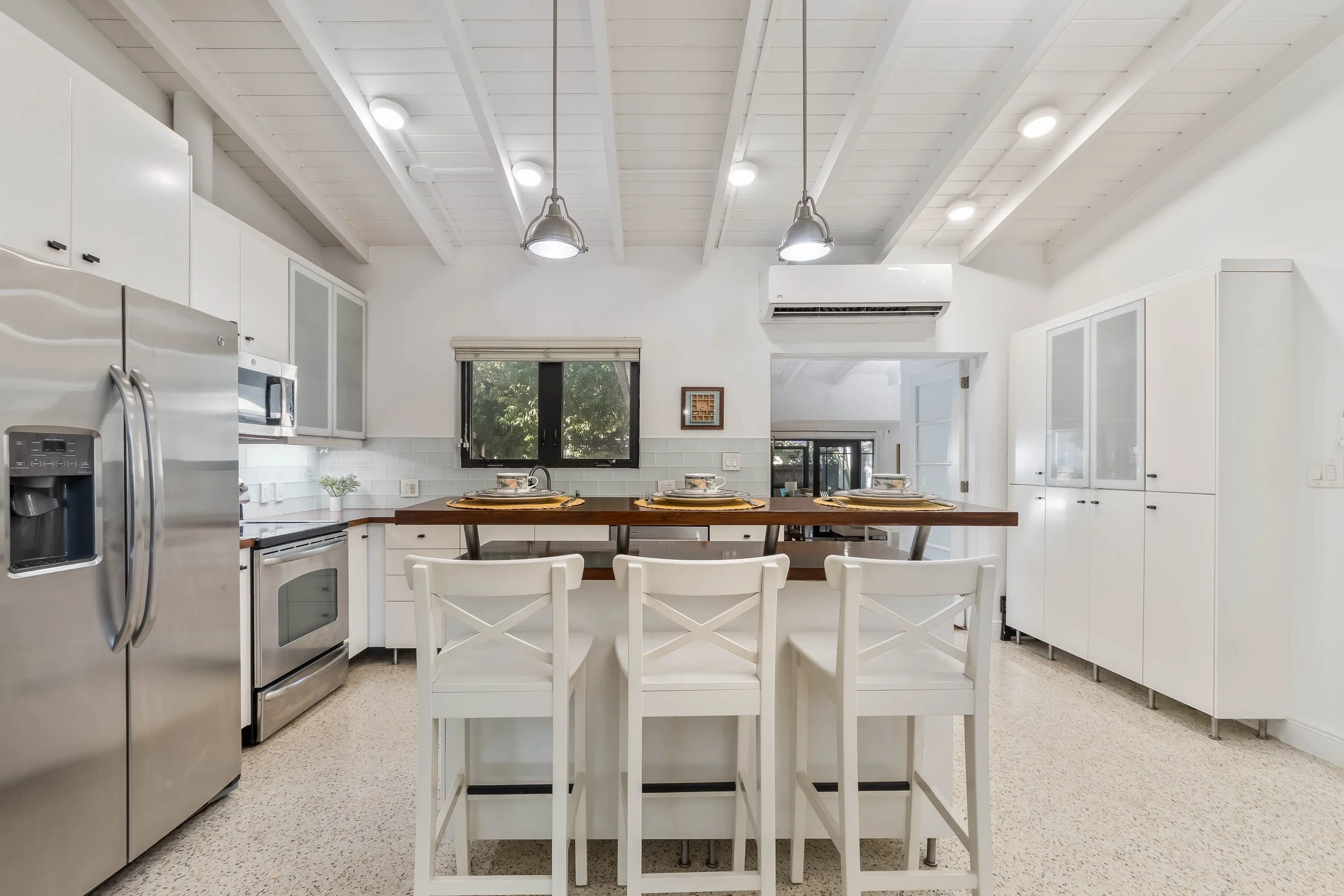 Modern kitchen with white cabinets, stainless steel appliances, a wooden kitchen island with four white chairs, and a small window over the sink, featuring a beige roller shade and greenery outside.