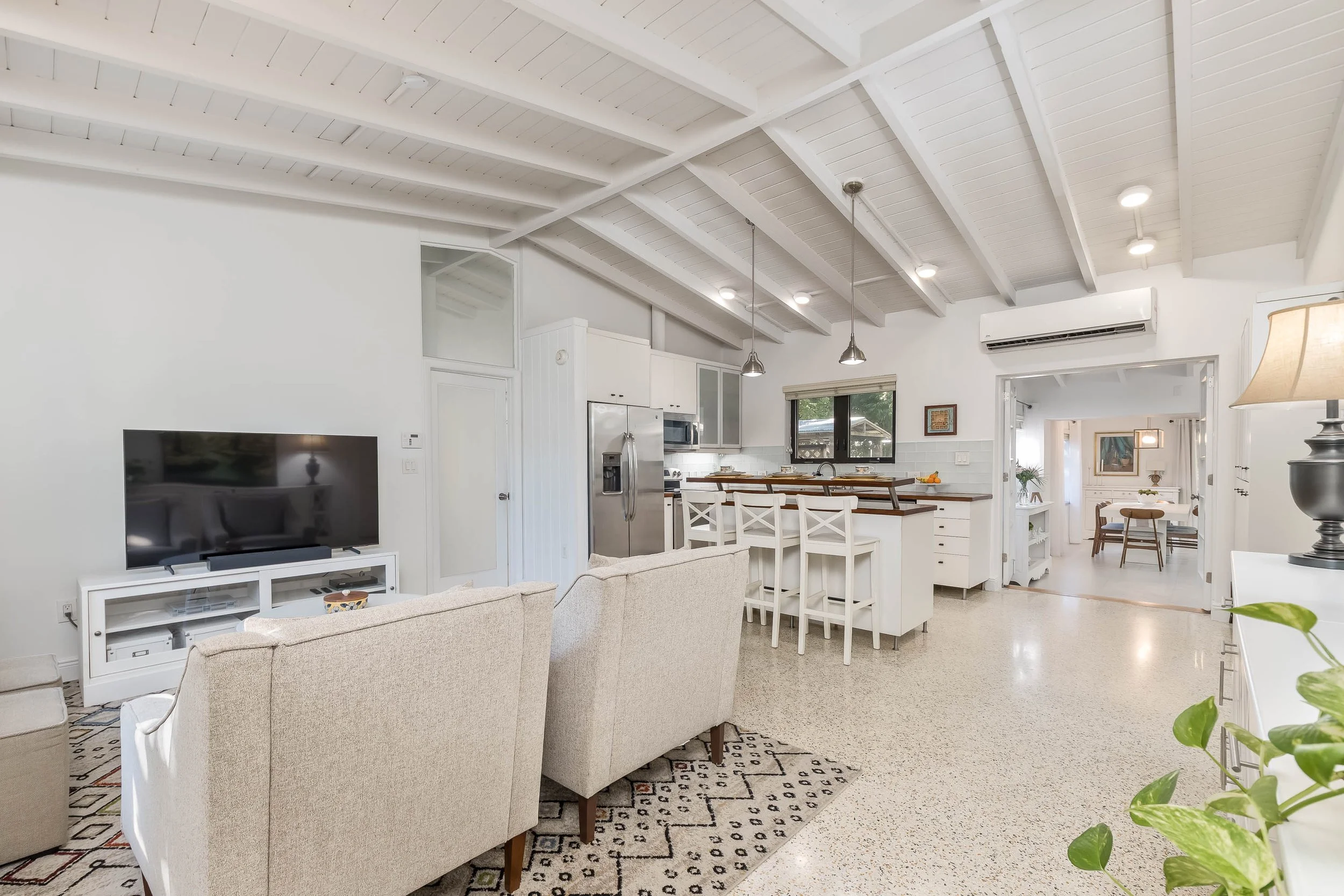 Open-concept living room and kitchen with white walls, exposed wooden ceiling beams, beige sofa, patterned area rug, flat-screen TV on a white TV stand, kitchen island with white chairs, stainless steel refrigerator, microwave, and appliances, large 
