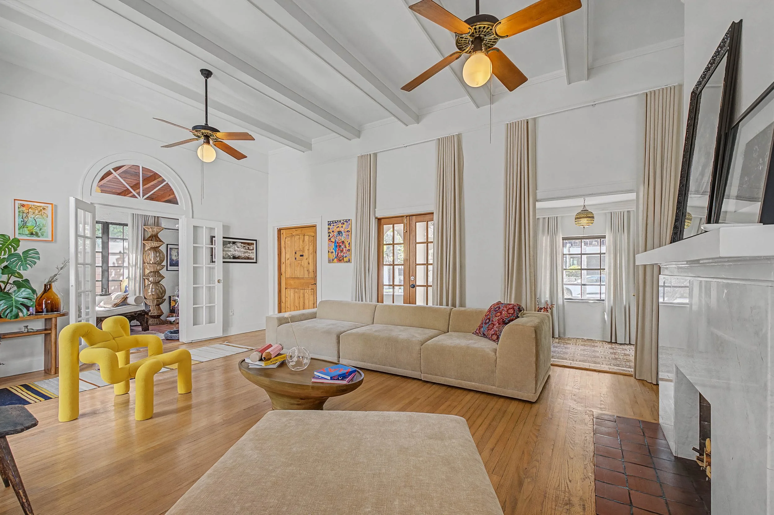 Living room with white walls, ceiling fans, beige sectional sofa, yellow accent chairs, wooden coffee table, hardwood flooring, windows with beige curtains, and doors leading to other rooms.