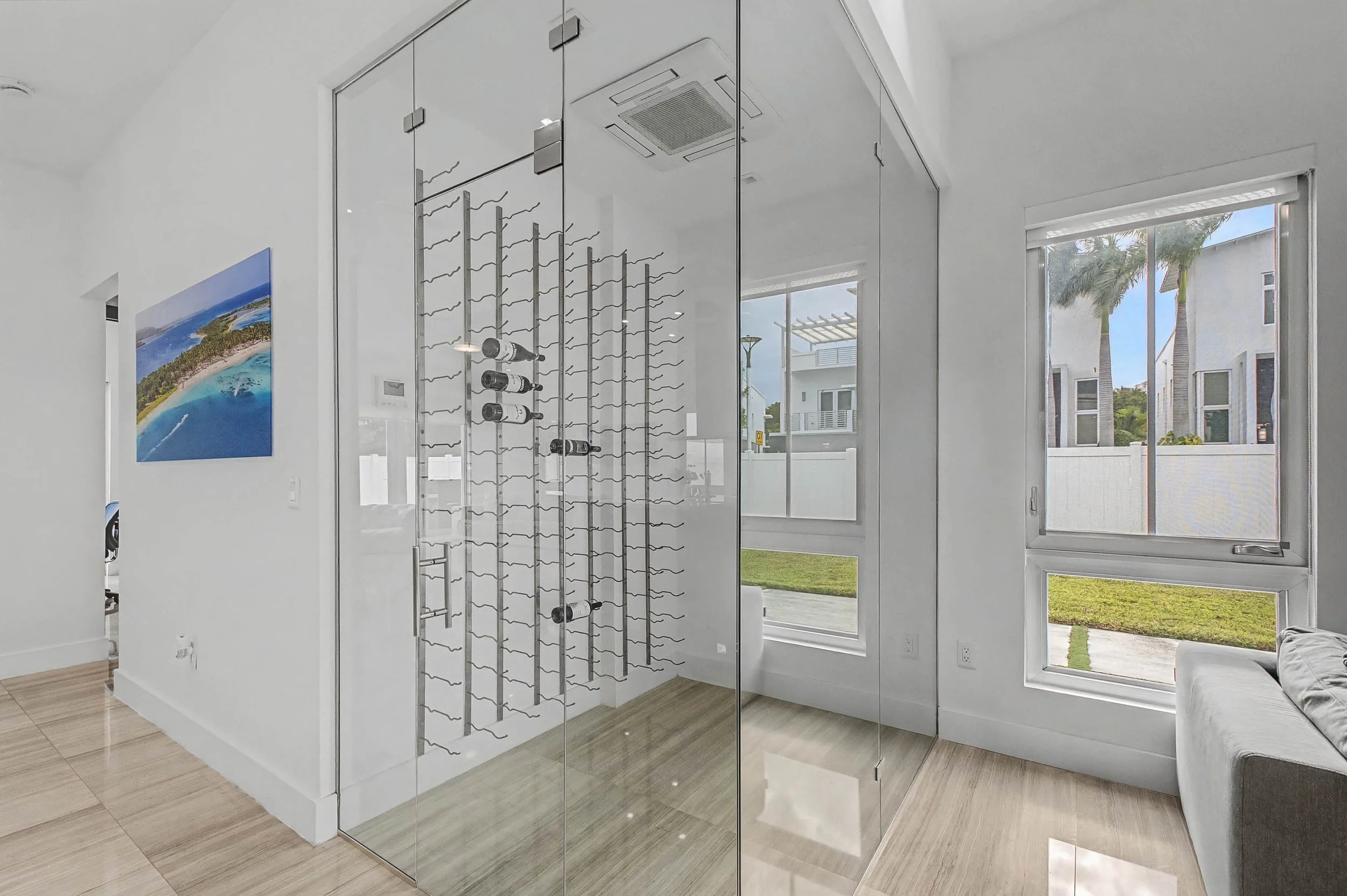 Interior of a modern house with white walls, a glass wine cellar, large windows showing palm trees and neighboring houses, and a gray couch.
