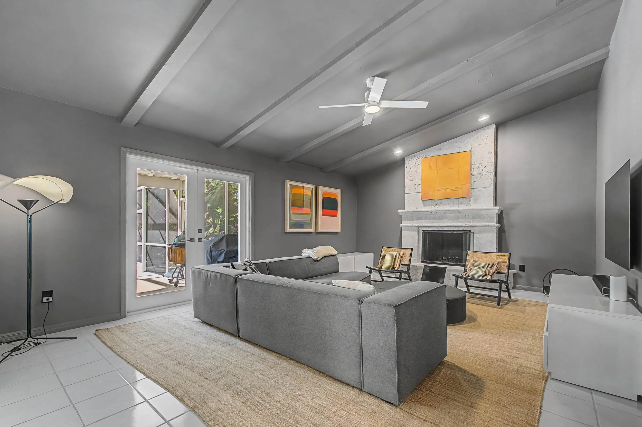 Living room with gray walls, a fireplace, and modern art on the walls. It has a large gray sectional sofa, two wooden chairs, and a white media console. There is a sliding glass door leading to an outdoor patio, a floor lamp, and a ceiling fan.