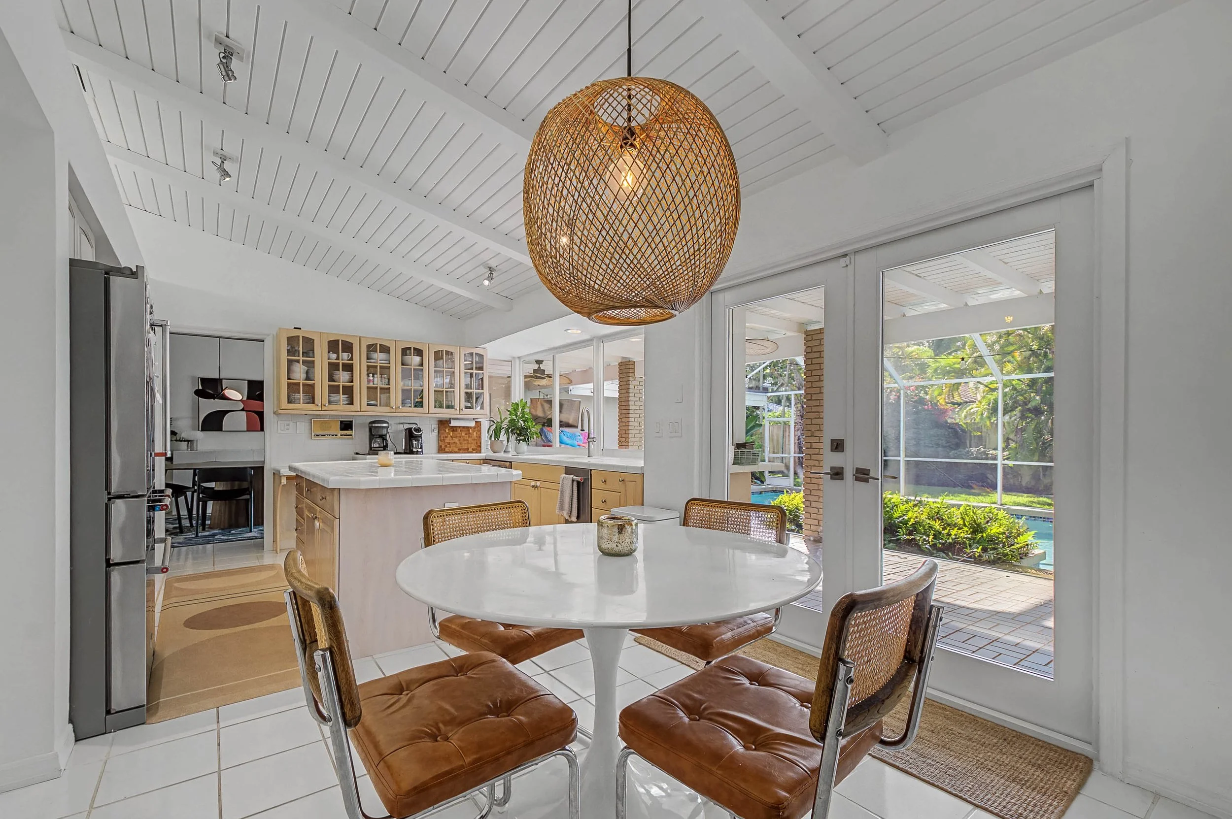 Dining area with a round white table and four vintage chairs with leather seats, a woven pendant light overhead, and a kitchen in the background with cabinets, a coffee maker, and a sliding glass door leading outside to a backyard with a pool.