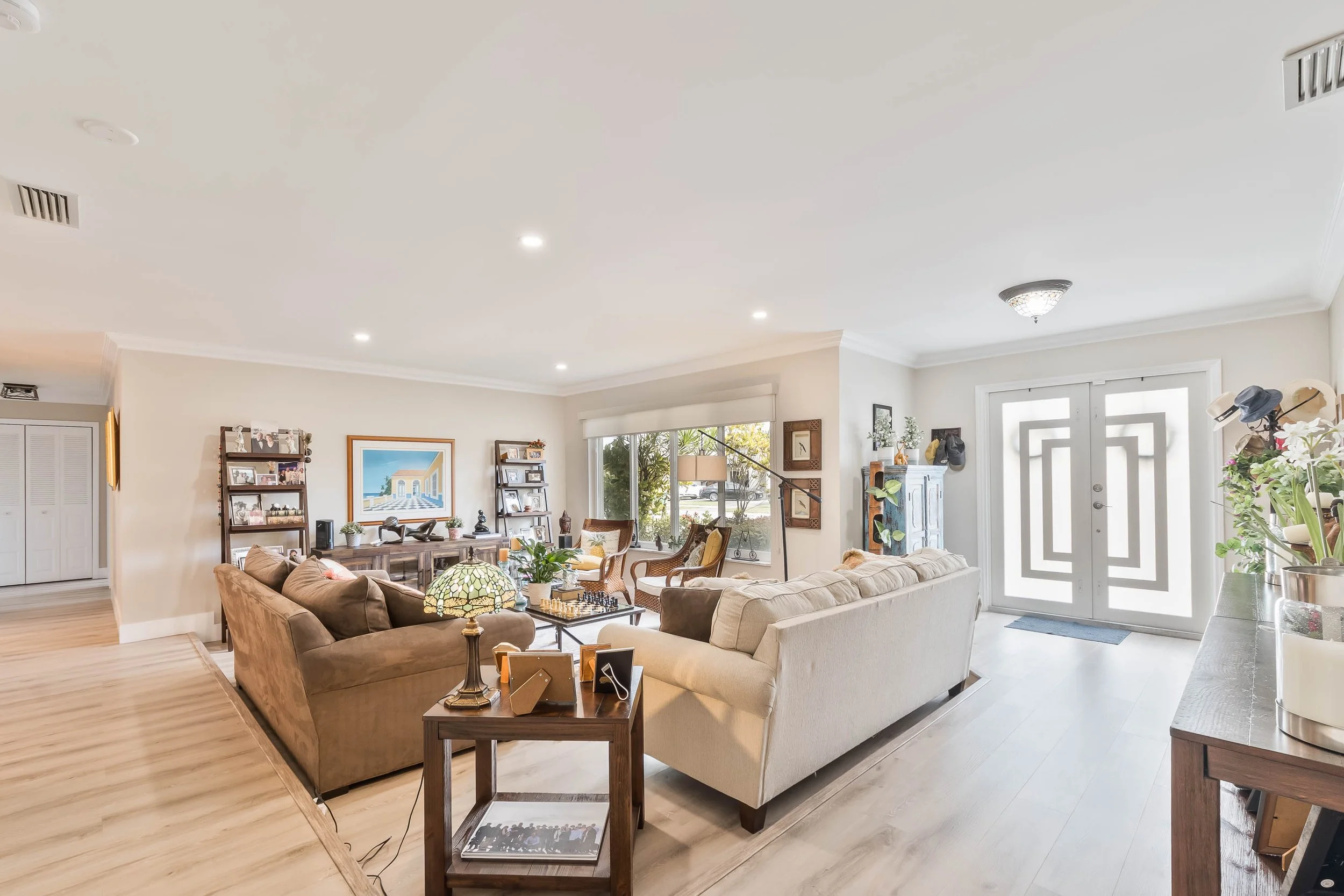 Bright living room with beige and brown couches, wood flooring, large window with blinds, and front door with geometric design.