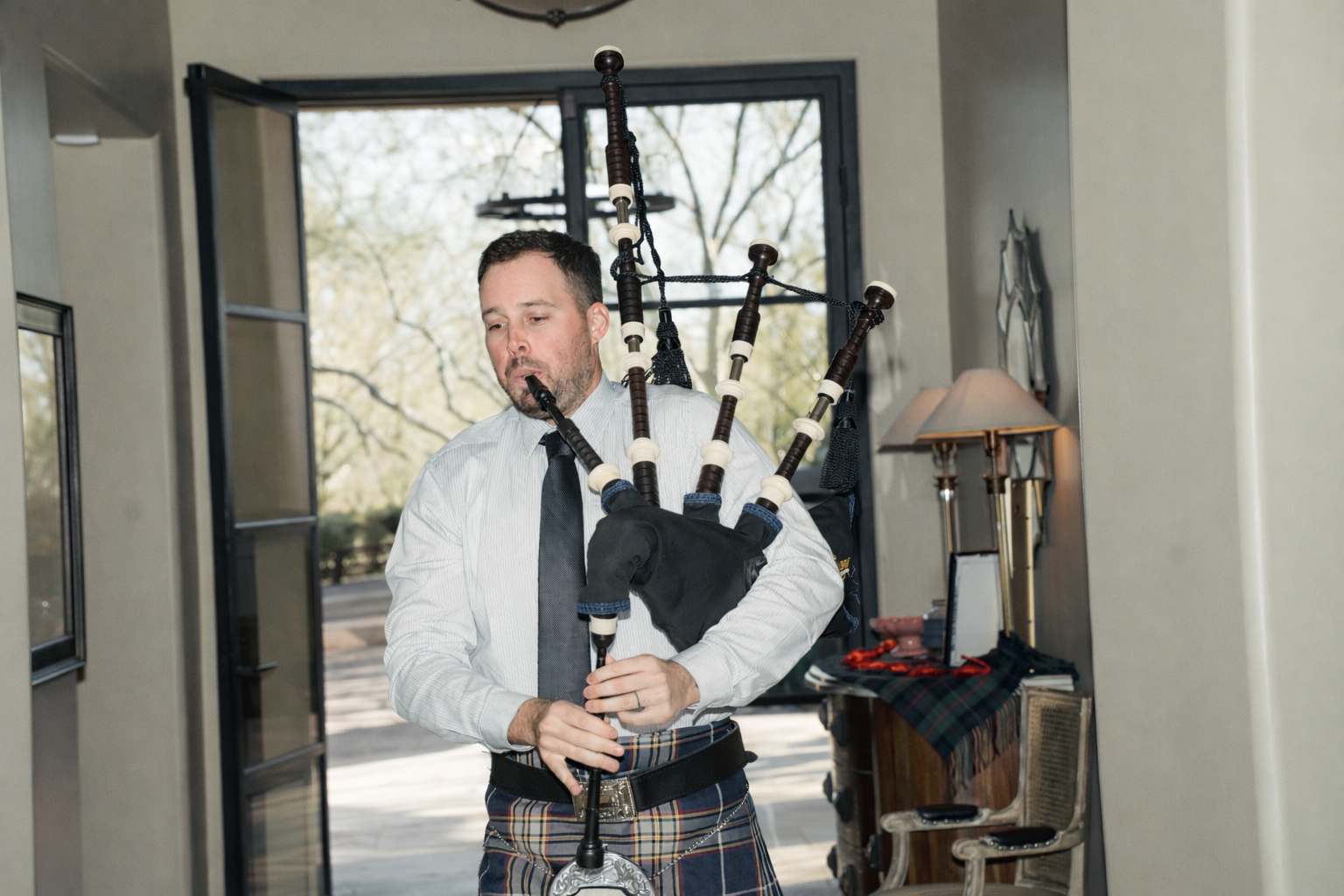 A man wearing a kilt is playing a set of bagpipes indoors near an open door, with trees visible outside.