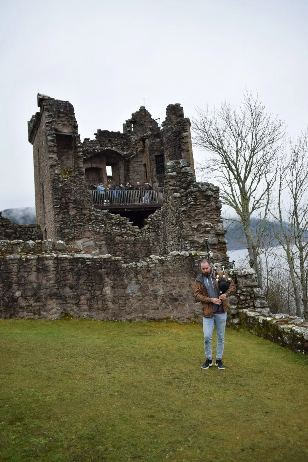 A man playing the bagpipes outdoors in front of a historic stone castle on a cloudy day.