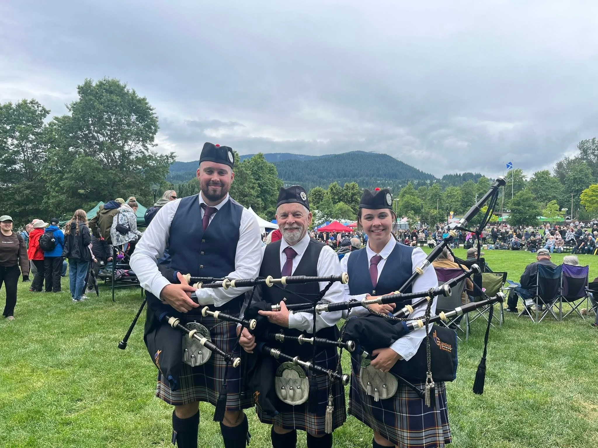 Three people in traditional Scottish attire holding bagpipes at an outdoor event with a crowd and mountains in the background.