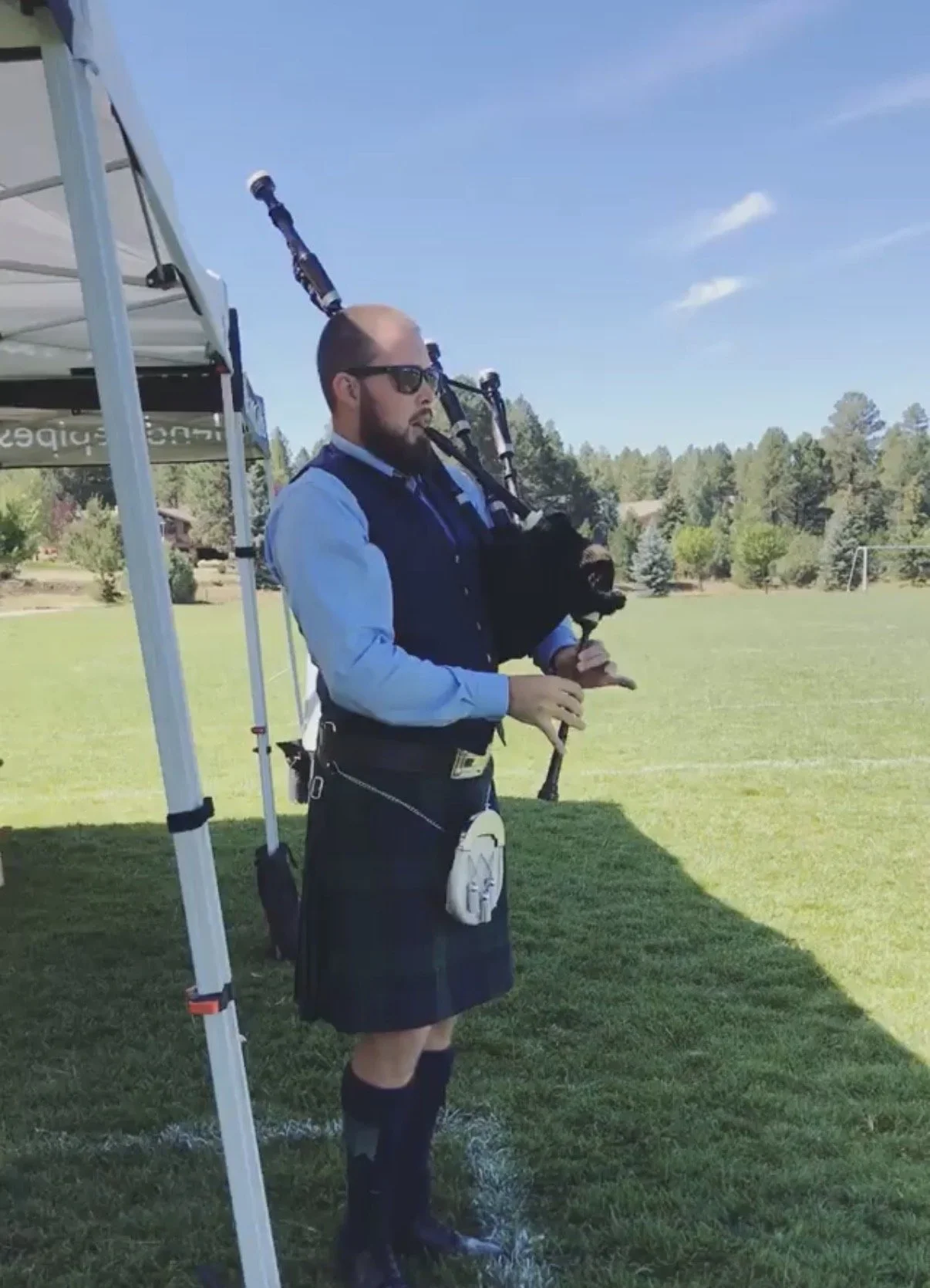 Man in uniform playing bagpipes outdoors on a grassy field under a blue sky.
