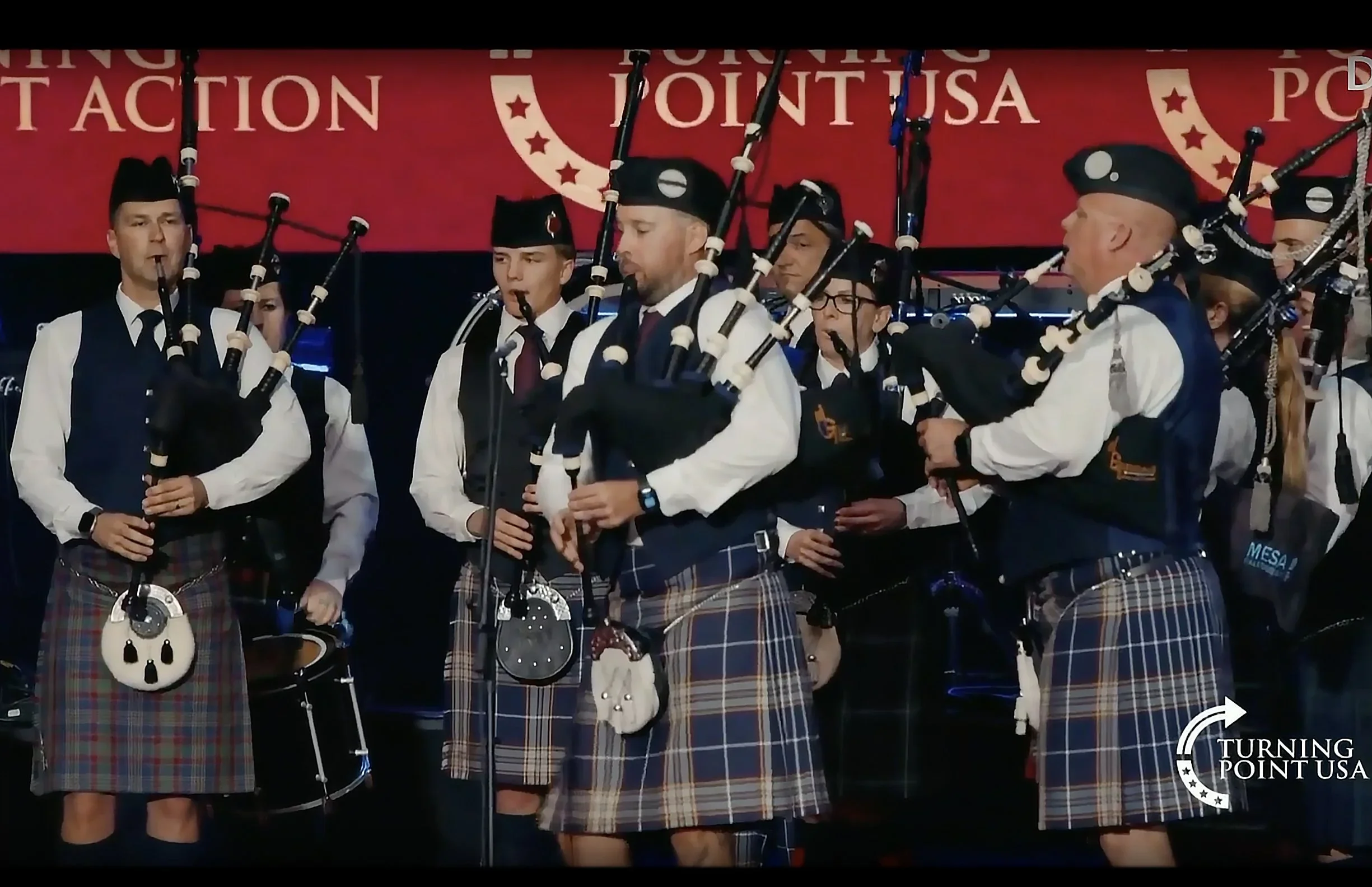 A group of musicians dressed in traditional Scottish attire, including kilts, playing bagpipes on stage during a performance.