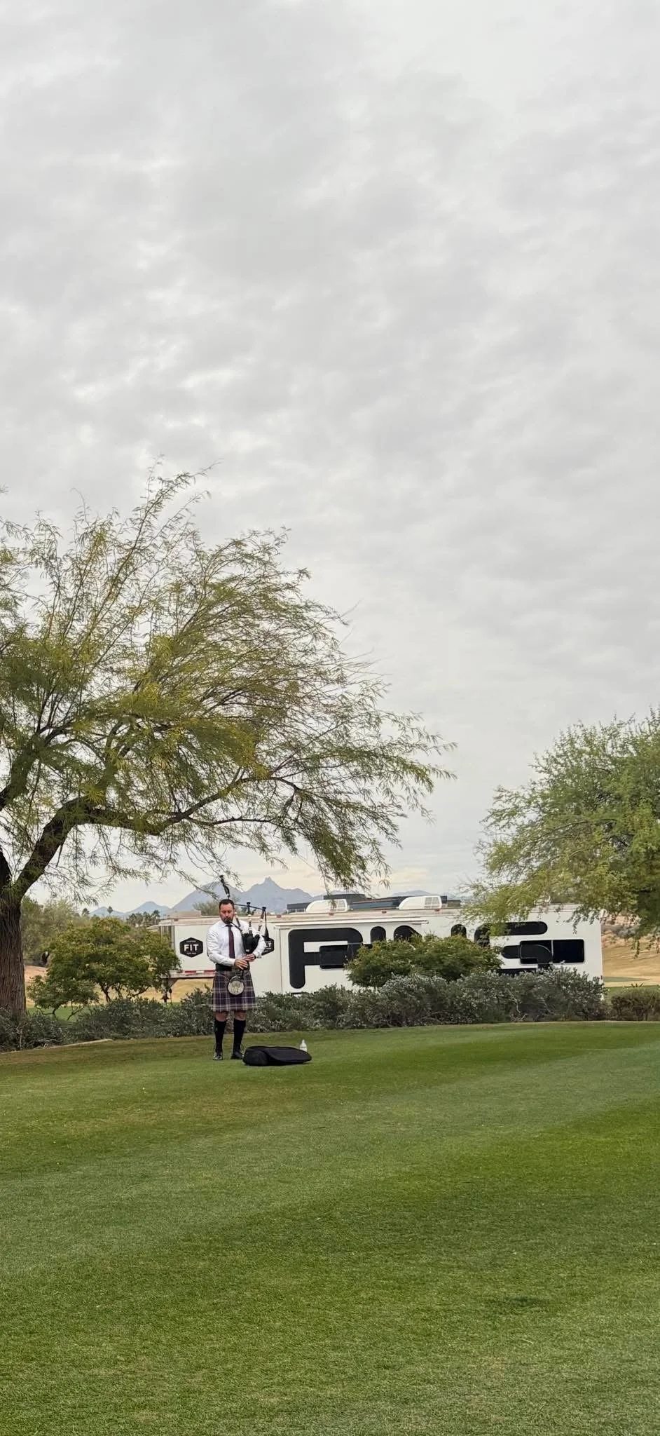 A man in a kilt playing bagpipes on a golf course with trees and mountains in the background, and a large trailer behind him.